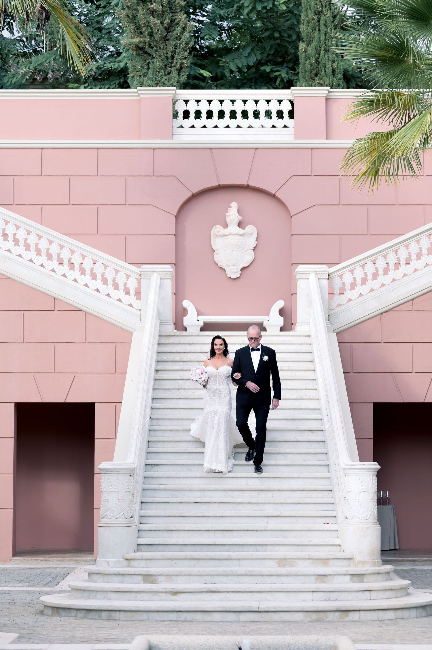 Bride in an off-shoulder white gown and groom in a black suit walk down white marble stairs at a pink palace wedding venue.