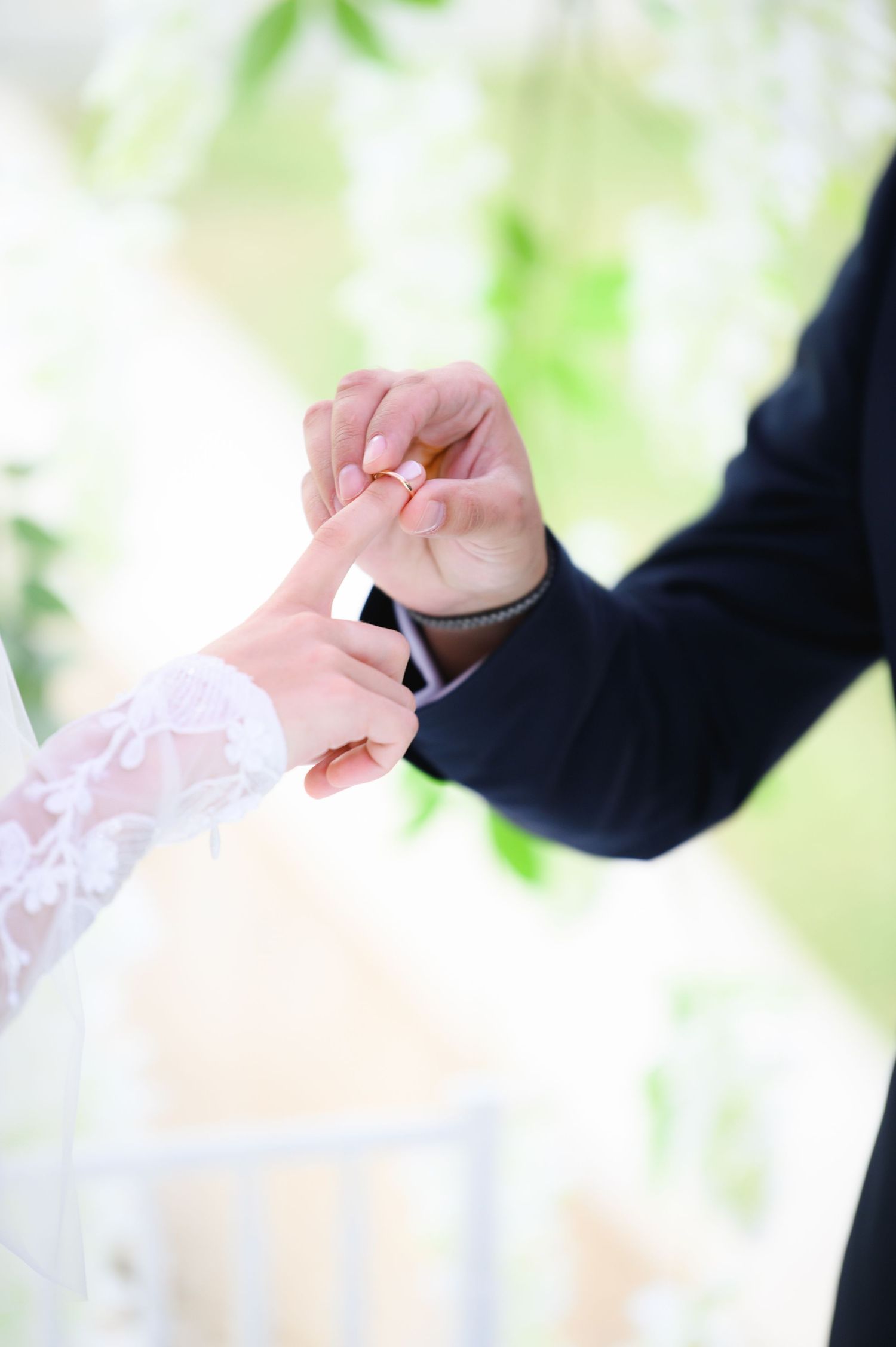 Groom in a dark suit slides a wedding ring onto the bride's finger during a ceremony, with soft white flowers in the background.