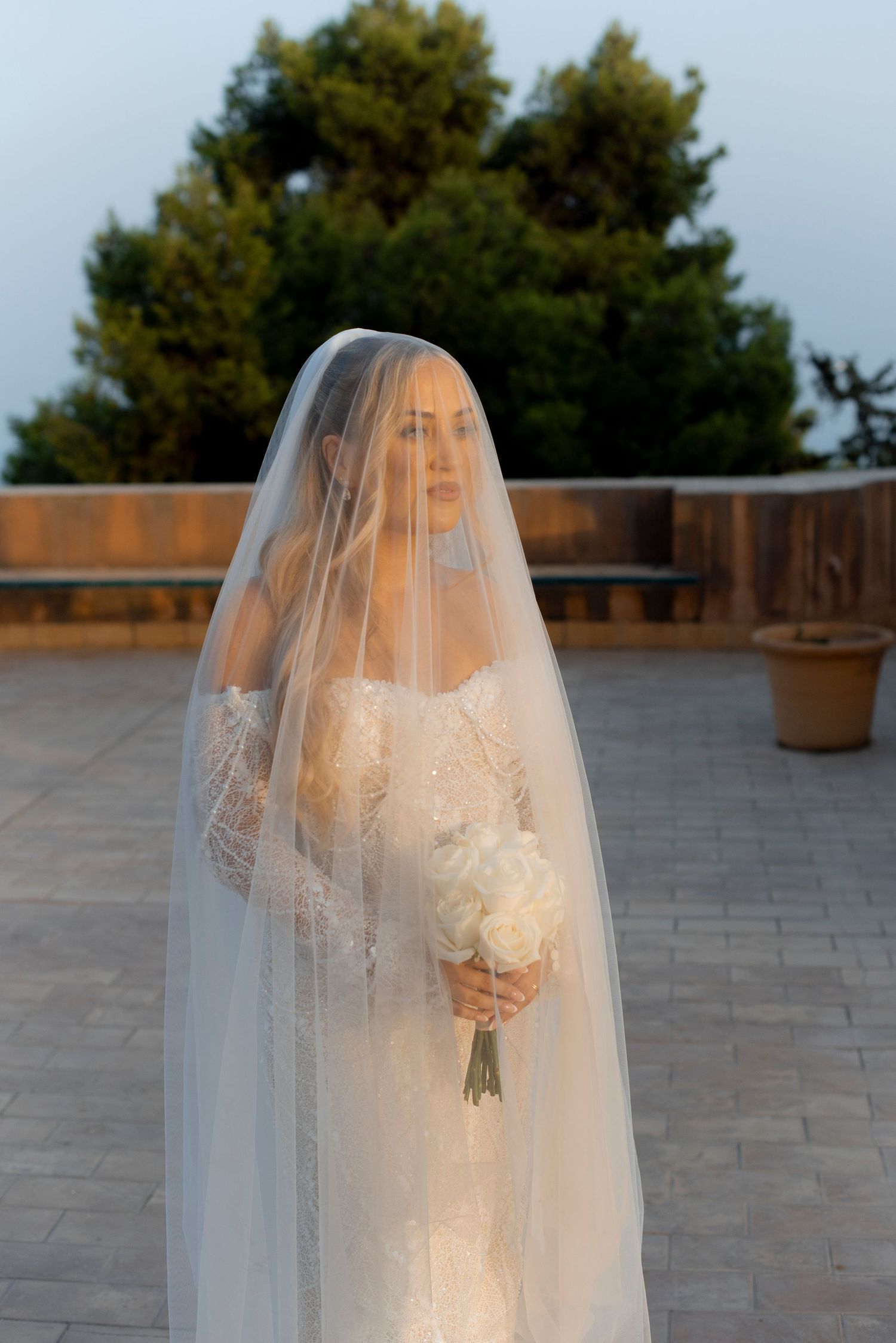 Bride in a lace wedding gown and long sheer veil holding a white floral bouquet on a stone terrace at sunset.
