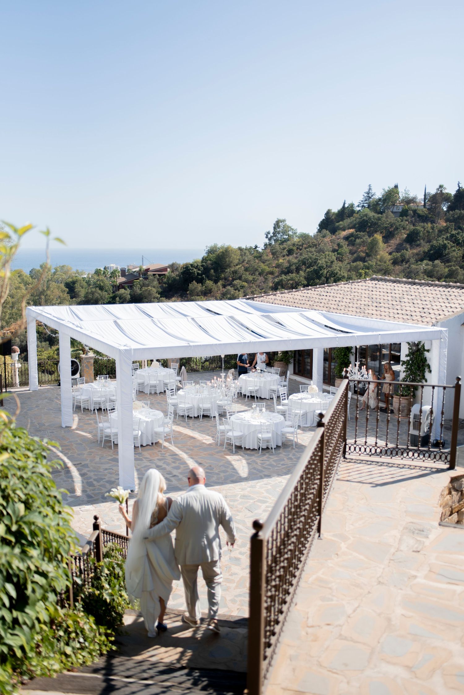 Bride and groom walk toward an outdoor wedding reception with white draped tables under a canopy overlooking green hills and the sea.