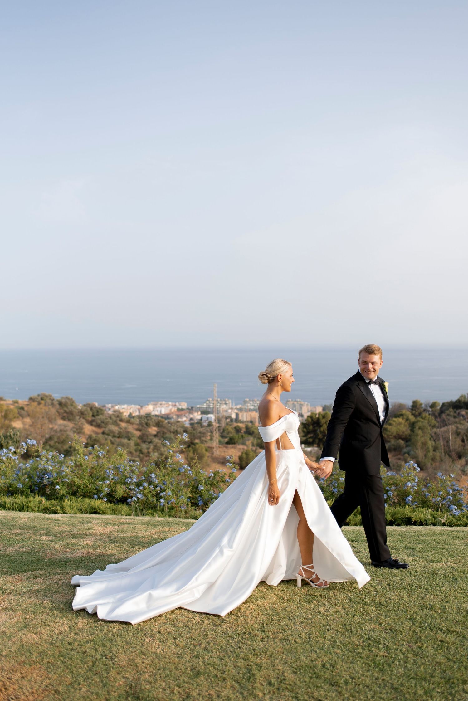 Bride in a white off-shoulder gown holds hands with groom in black tuxedo while walking on a grassy hillside above a coastal town and sea.
