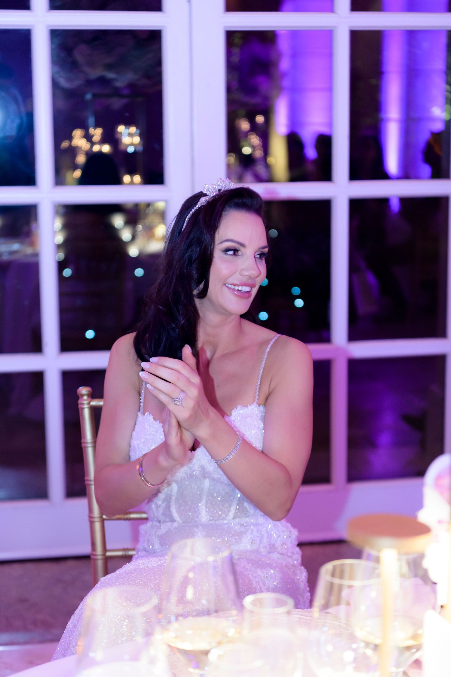 Smiling bride in a tiara and sparkling wedding dress claps at her reception table with purple lighting and wine glasses.