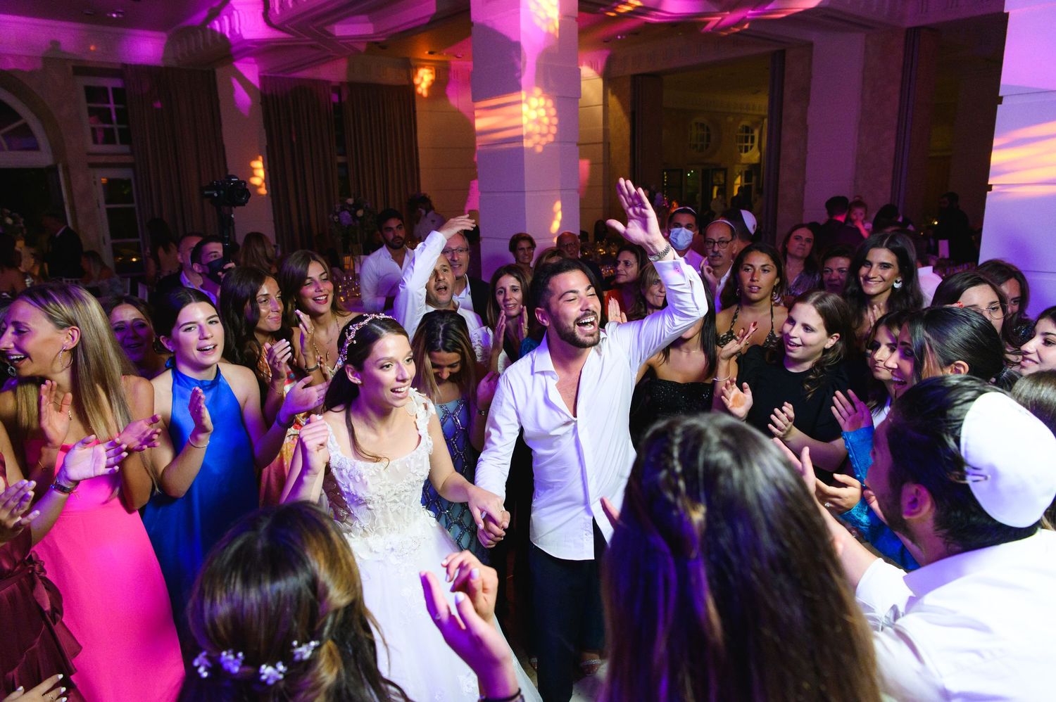 Groom in a white shirt energizes a crowd of dancing wedding guests under colorful lights at the reception.