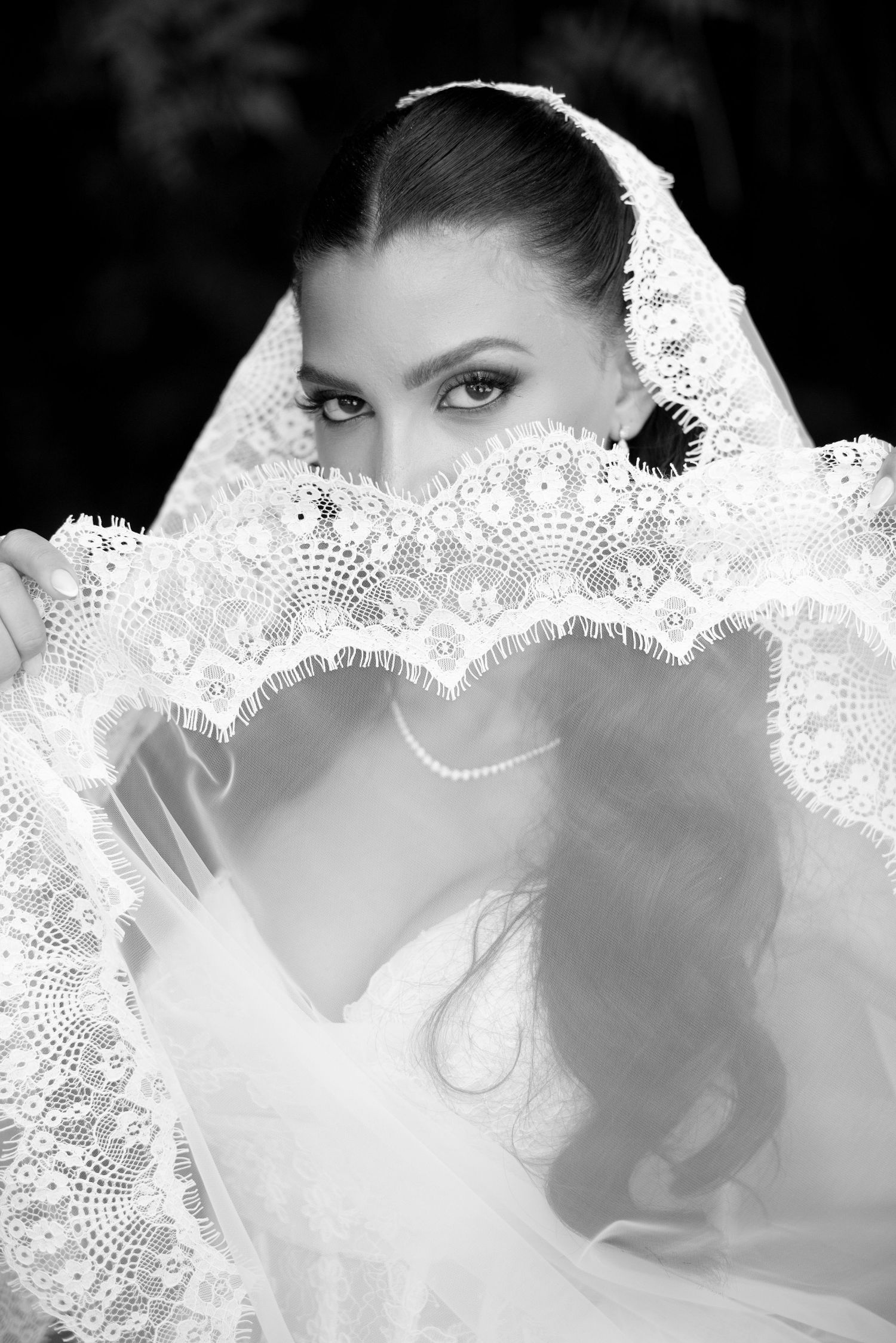 Black and white portrait of a bride peeking over a delicate lace veil, wearing a pearl necklace and soft waves.