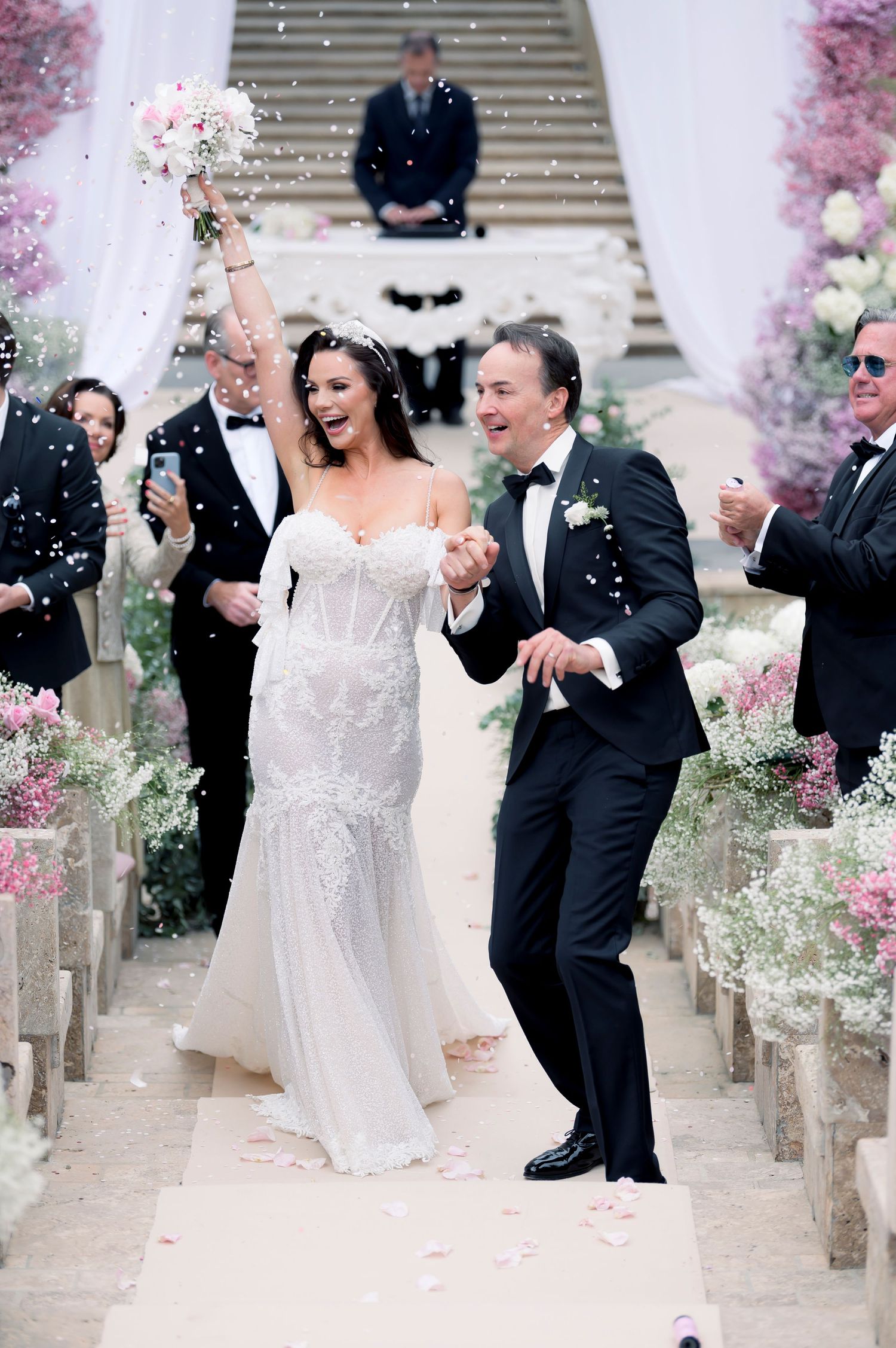 Bride in a lace gown and groom in a black tuxedo walk hand in hand down a petal-strewn aisle at a floral wedding ceremony.