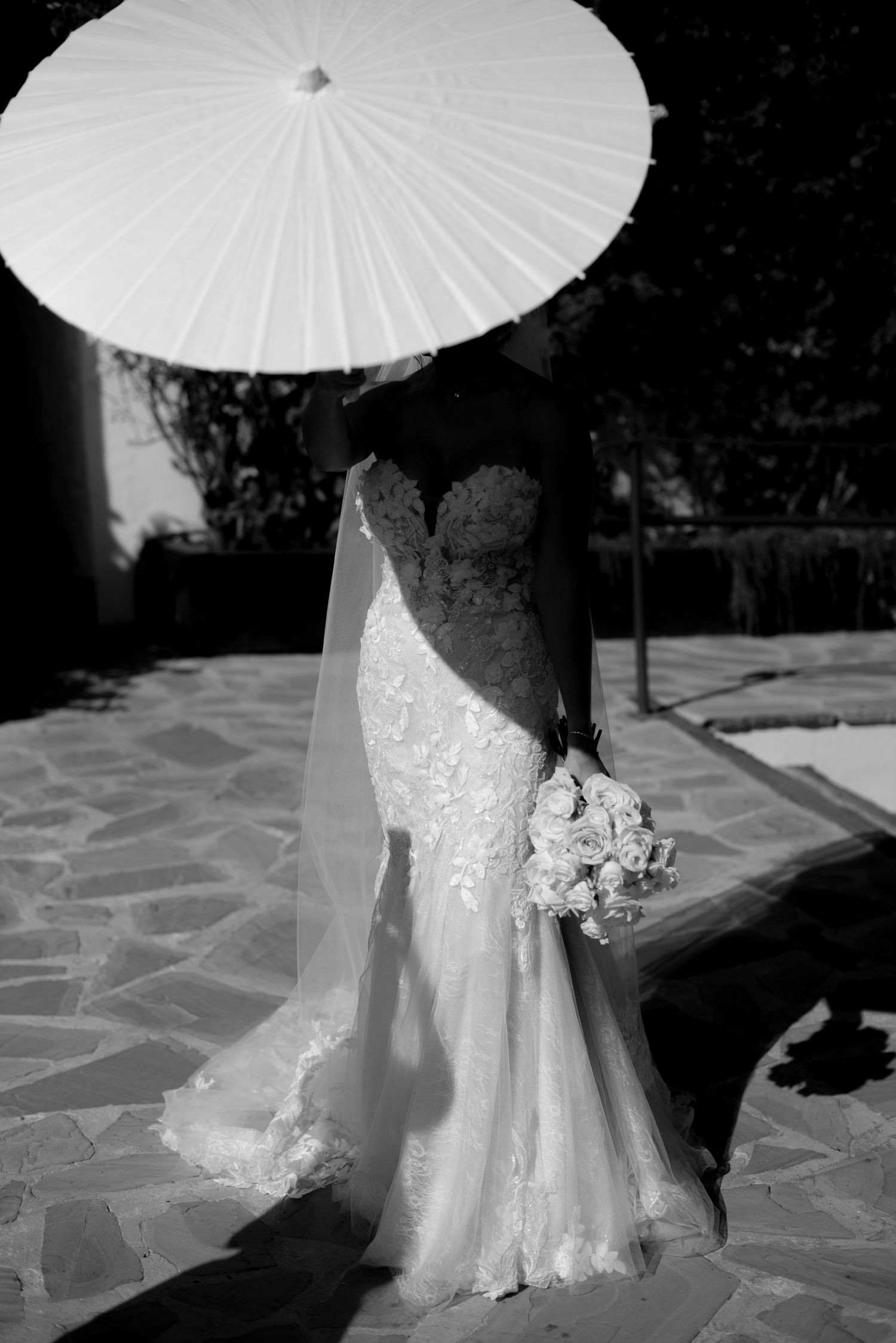 Black and white photo of a bride in a lace wedding gown and veil holding a bouquet under a paper parasol on a stone patio.
