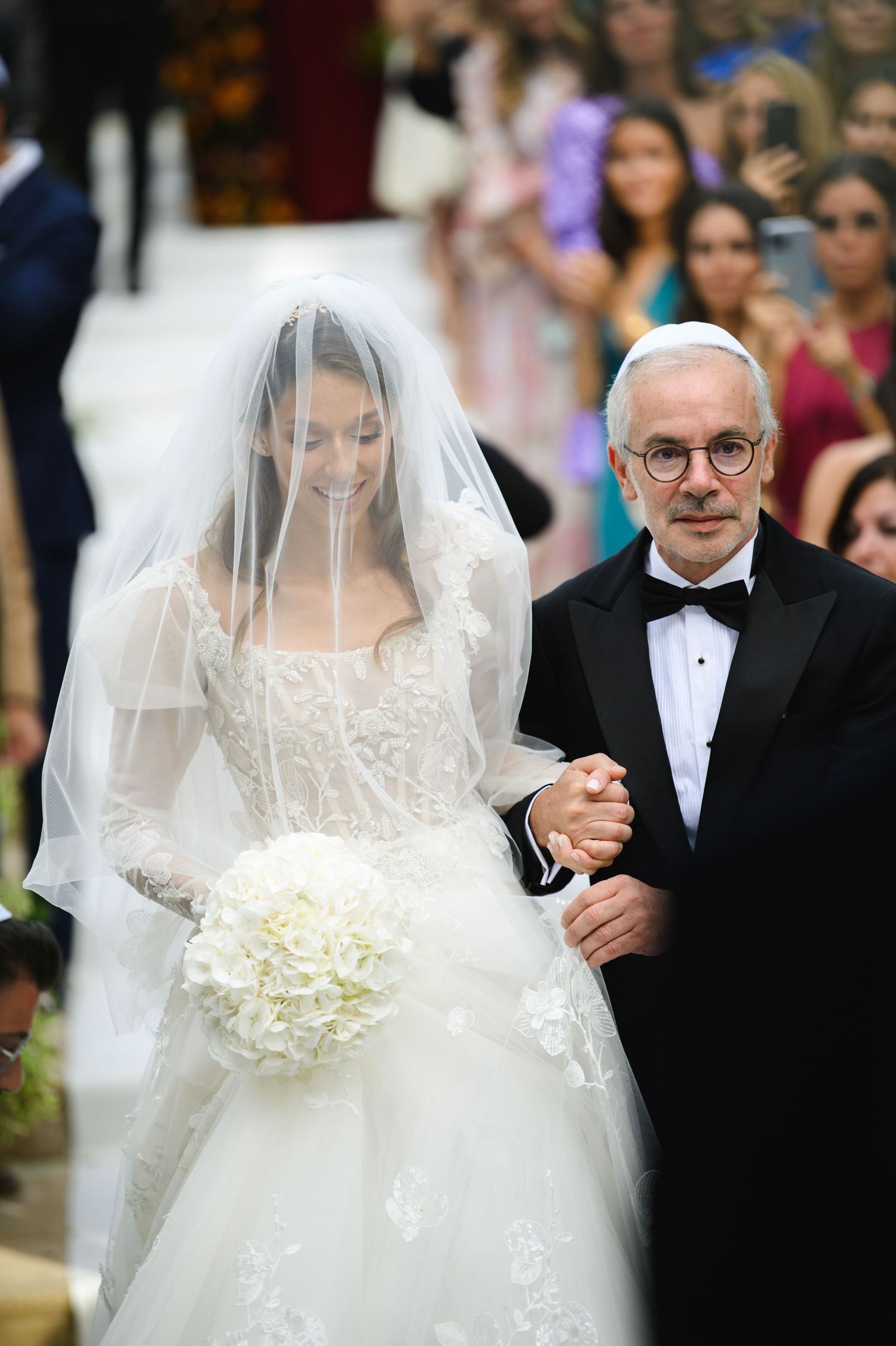 Father in a black tuxedo and kippah walks the veiled bride holding a white bouquet down the aisle.