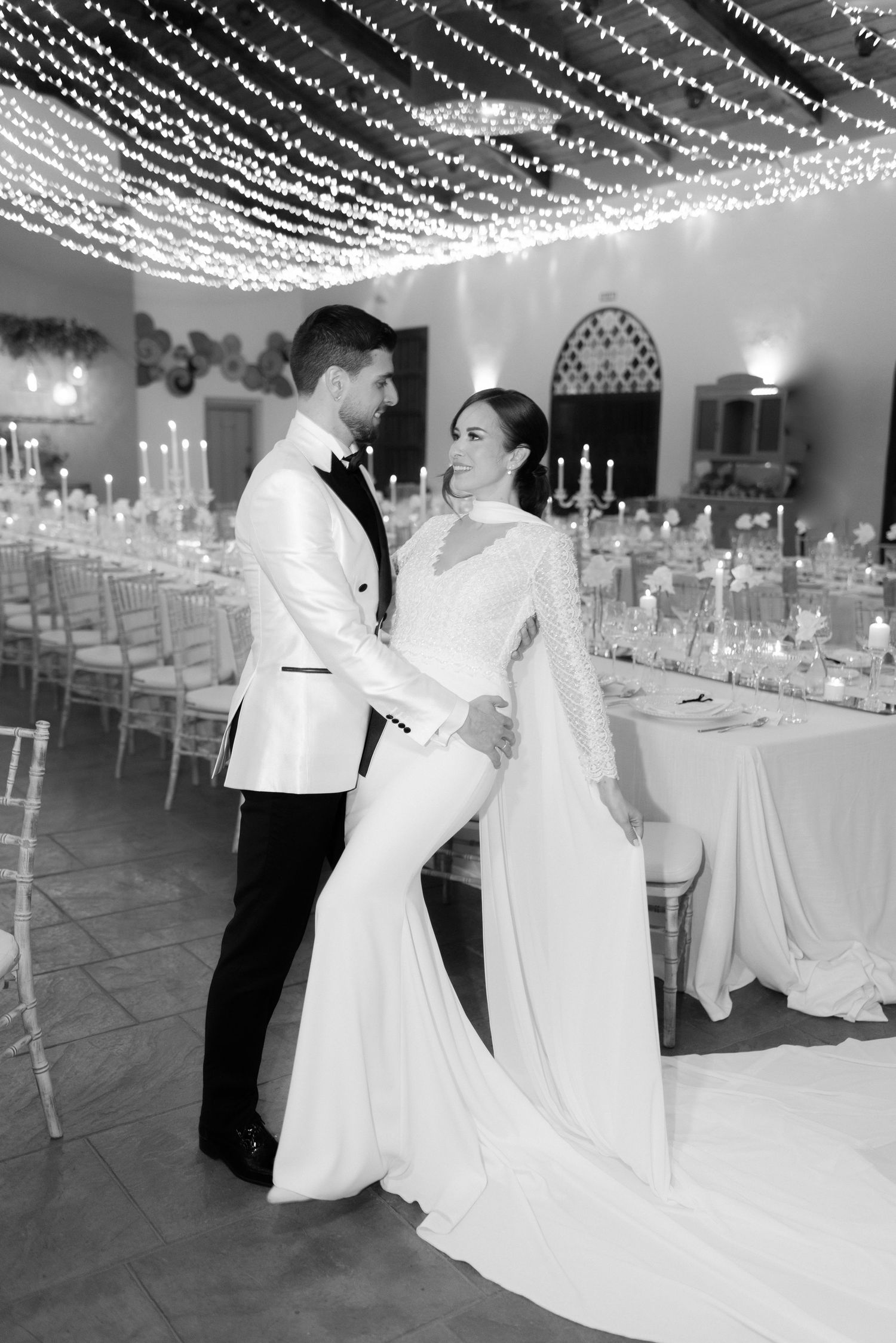Black and white photo of a bride and groom embracing in an elegant reception hall with string lights overhead and candlelit tables.