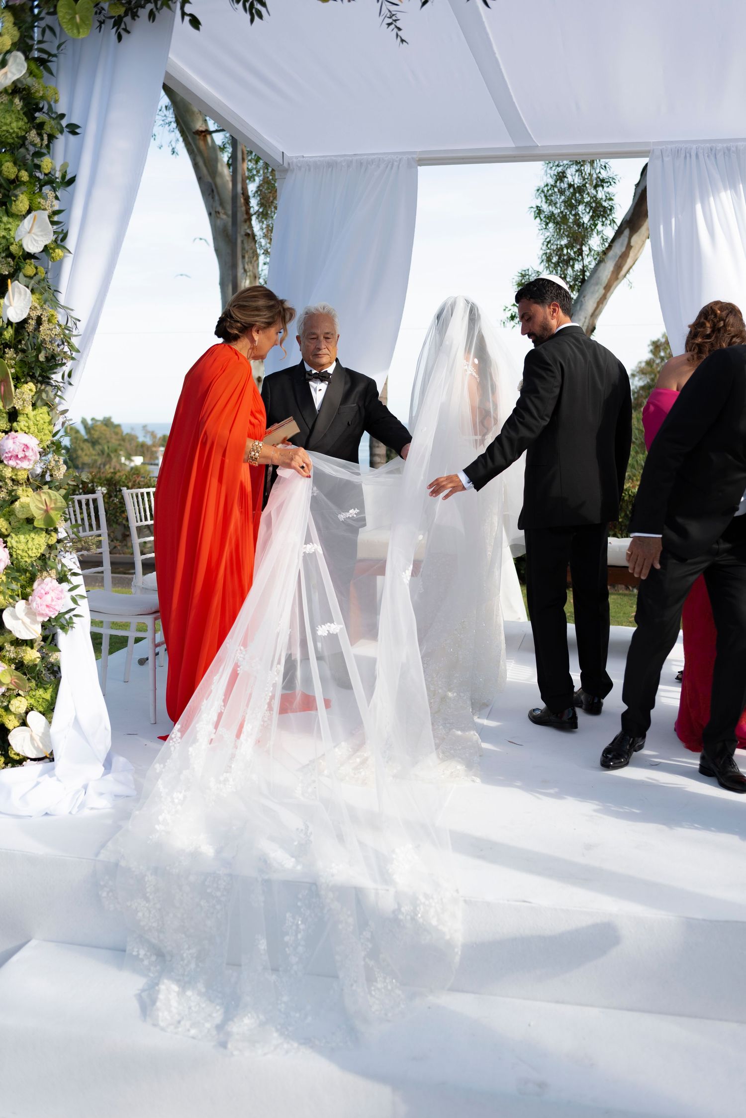 Bride in a long lace veil stands beneath a white outdoor wedding canopy as family in formal attire gather around her during the ceremony.