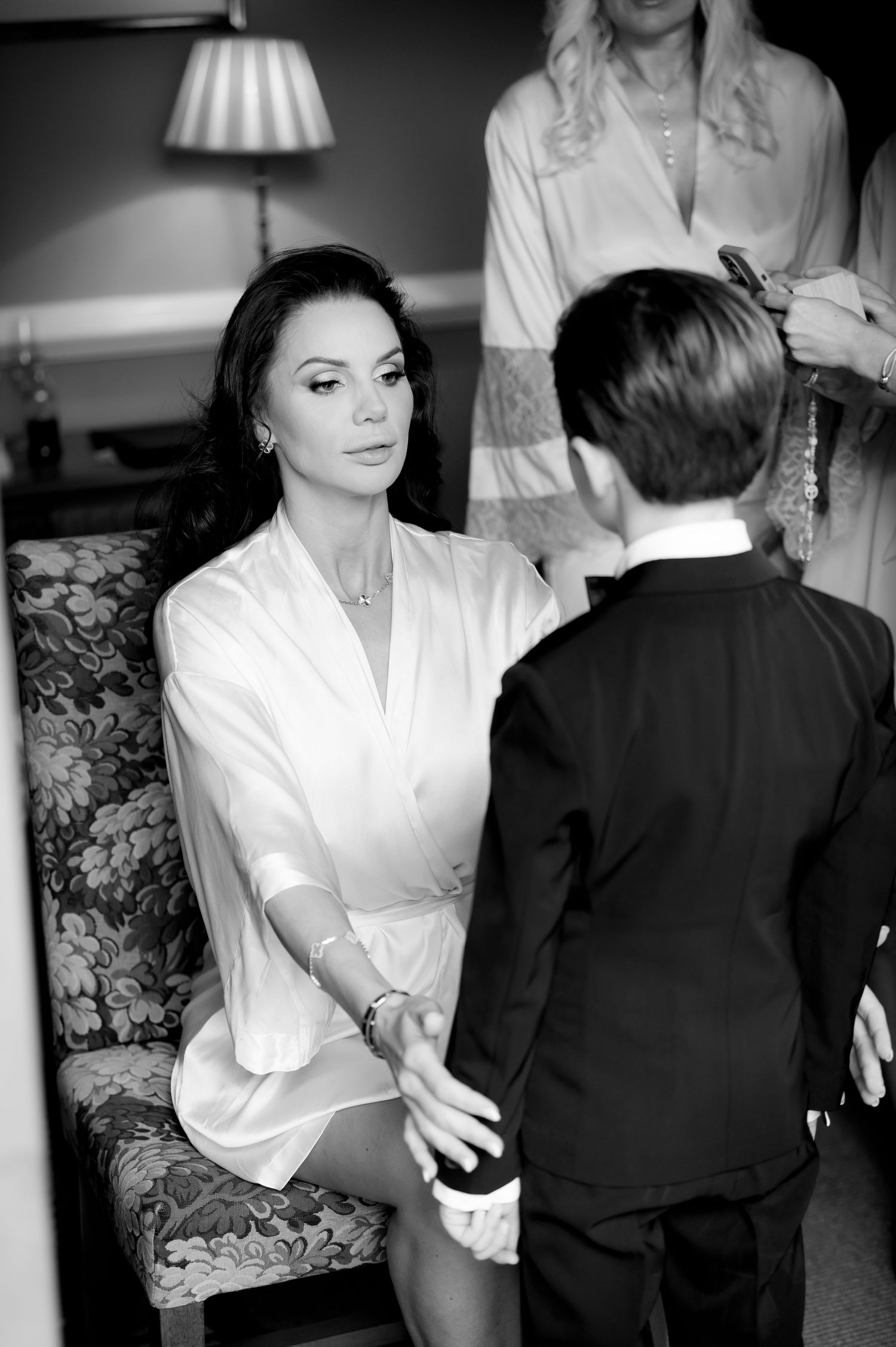 Black and white photo of a woman in a silk robe seated on a floral chair holding hands with a boy in a tuxedo before a wedding.