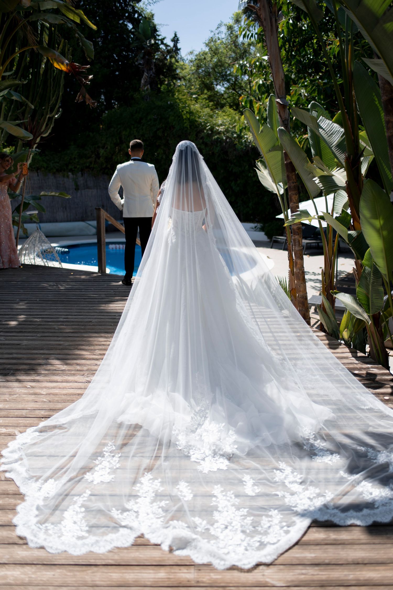 Bride in a lace wedding dress with a long sheer cathedral veil faces the groom in a white jacket on a wooden deck by a pool.