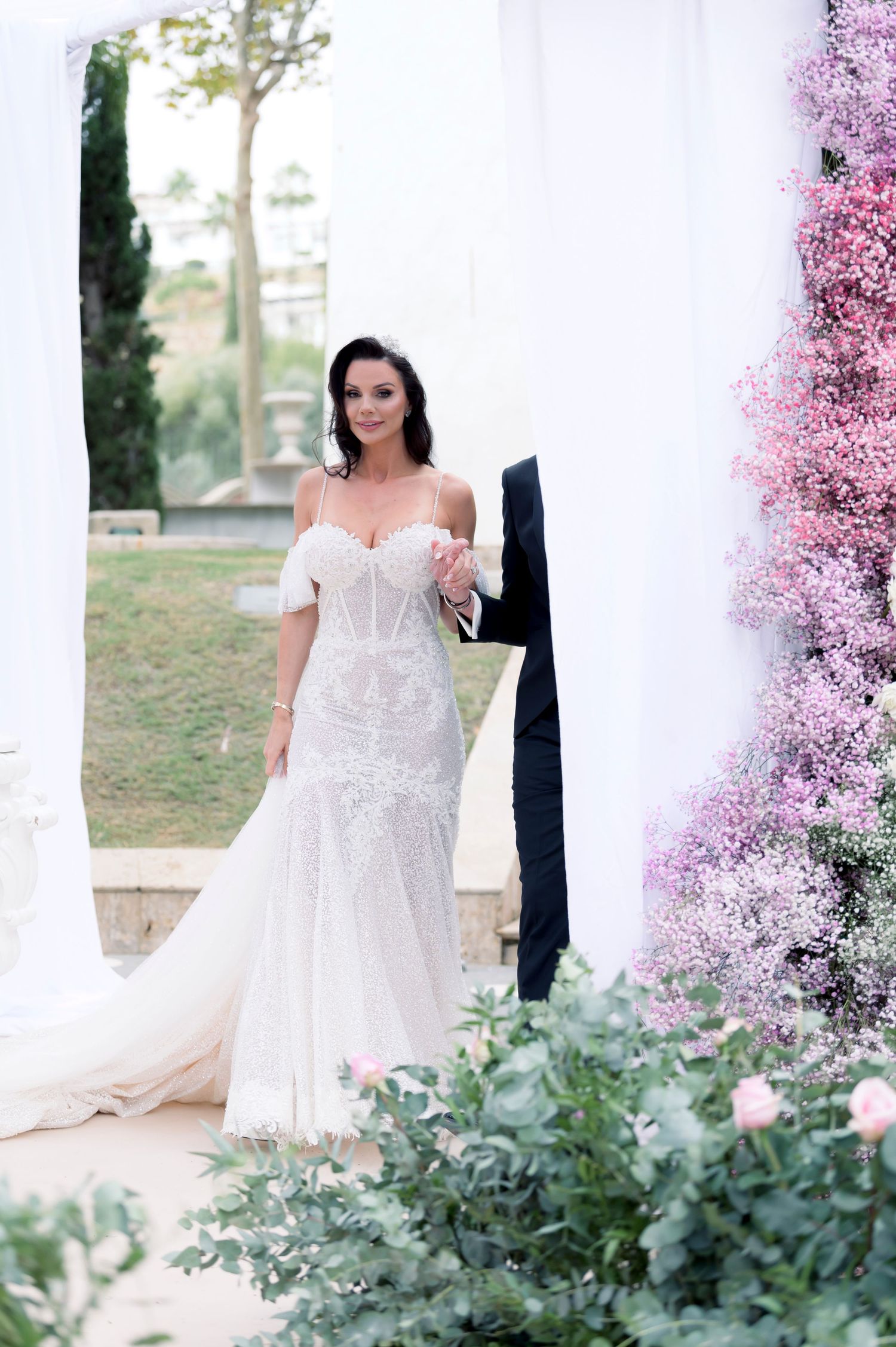 Bride in an off-the-shoulder white lace mermaid wedding gown walks beside a groom near a white drape and pink floral arrangement.