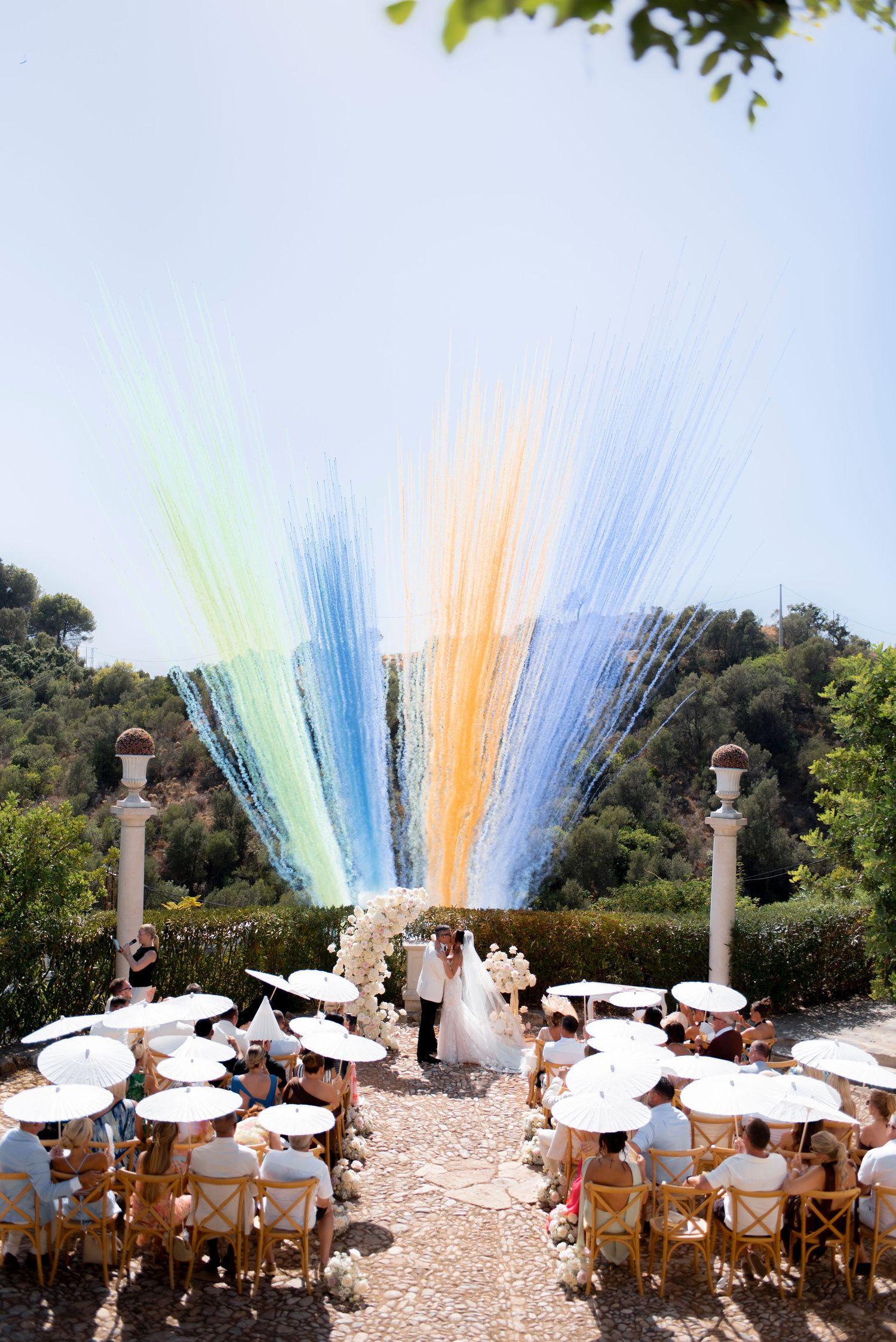 Outdoor wedding ceremony with a bride and groom beneath a rainbow of hanging ribbons, guests seated under white parasols in a garden.