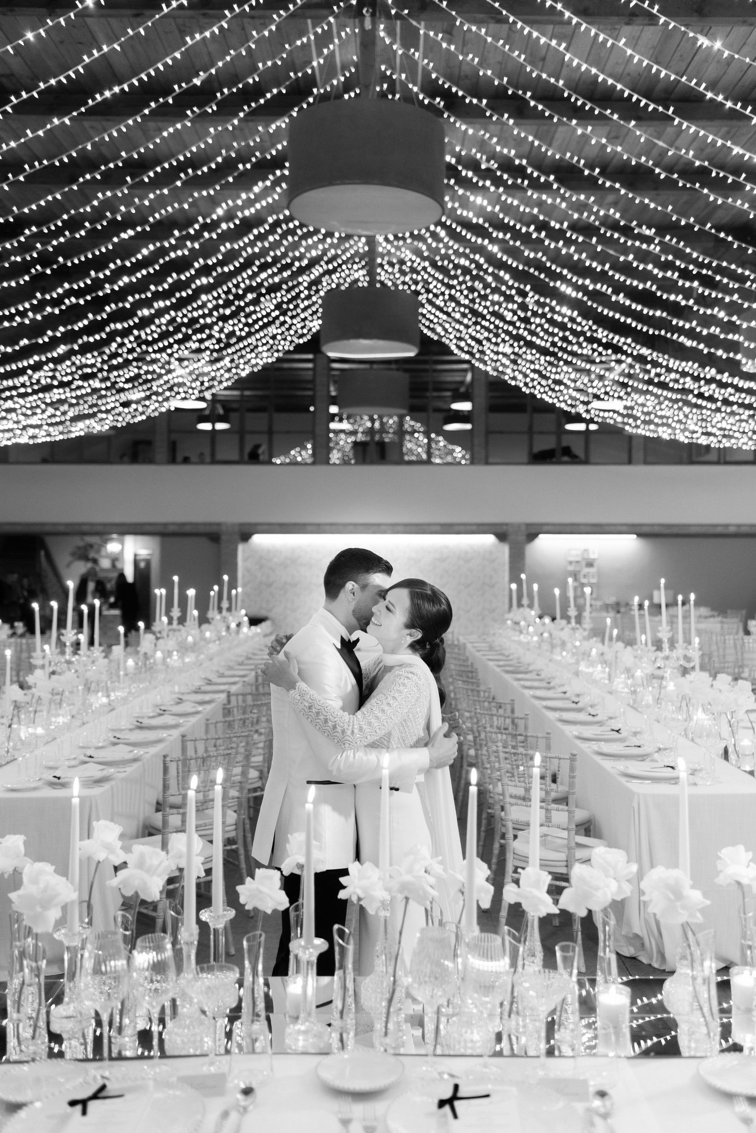 Bride and groom embrace between long banquet tables with candles, white flowers and canopy string lights at a wedding reception.