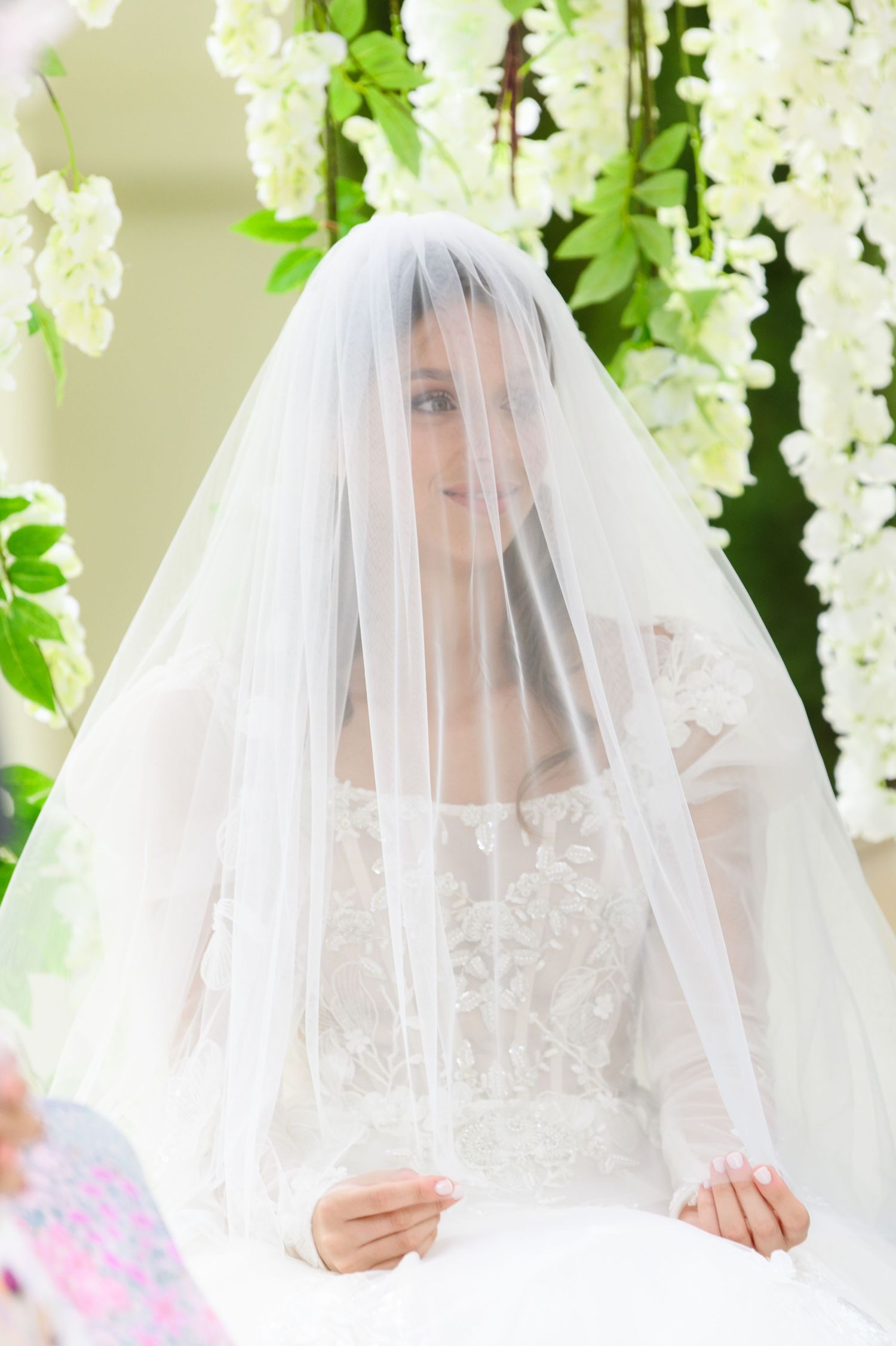 Bride in a lace wedding gown with a sheer veil covering her face, smiling under a floral arch of white blossoms.