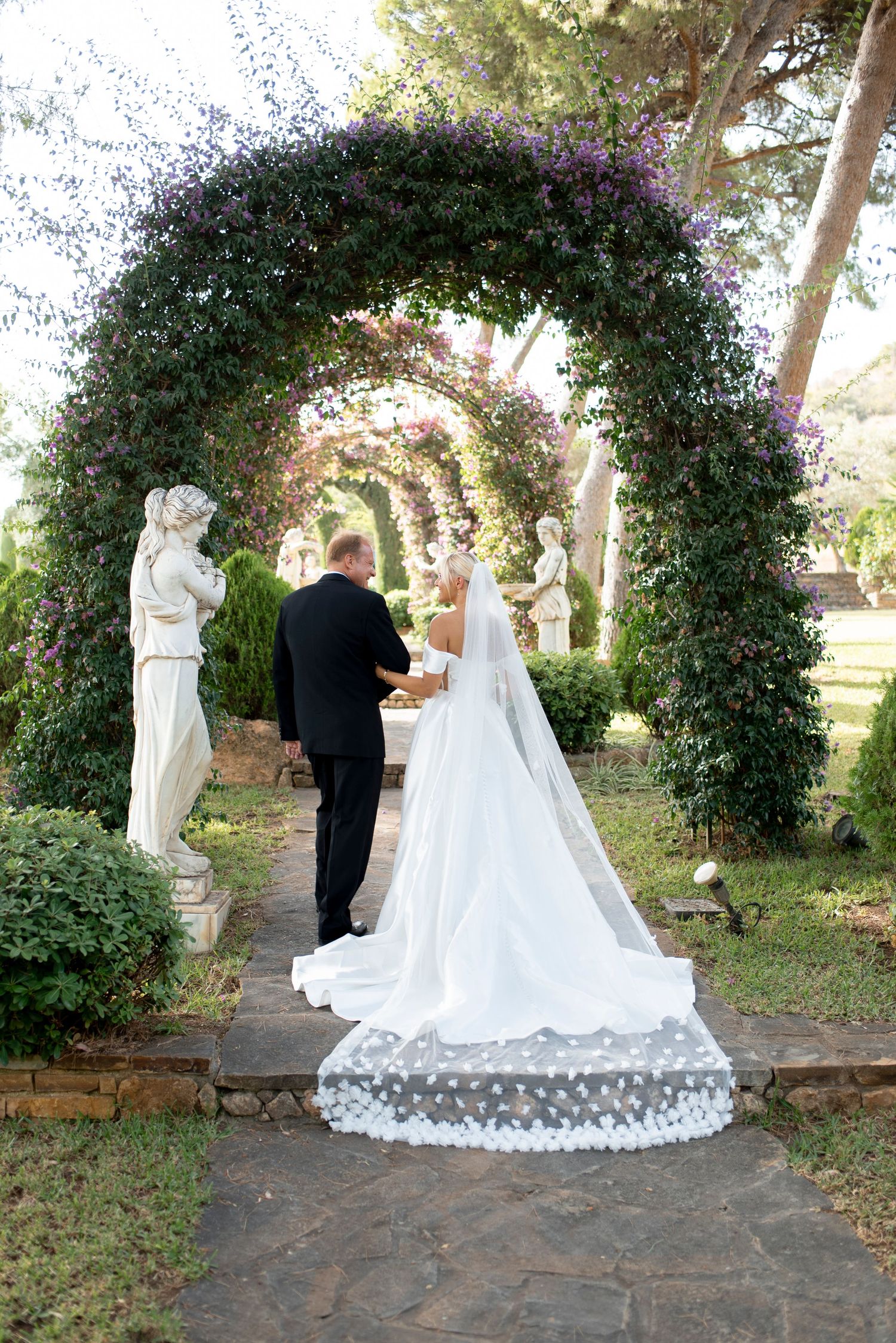 Bride in a long white gown and cathedral veil walks with her groom beneath a purple flowering garden arch flanked by marble statues.