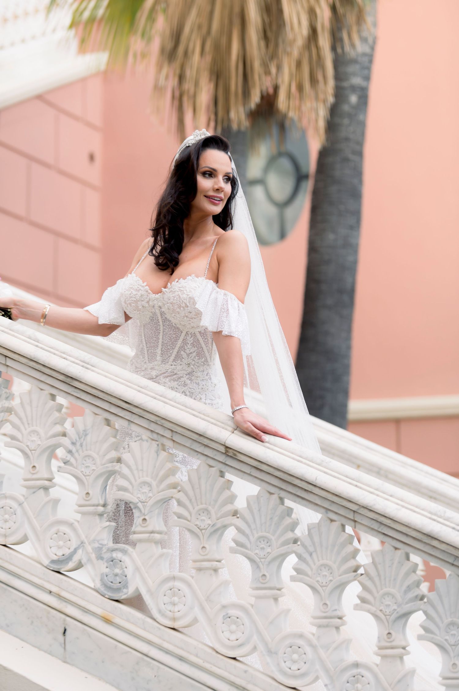 Smiling bride in an off-shoulder lace wedding dress and veil stands on an ornate white balustrade staircase beside a palm tree.