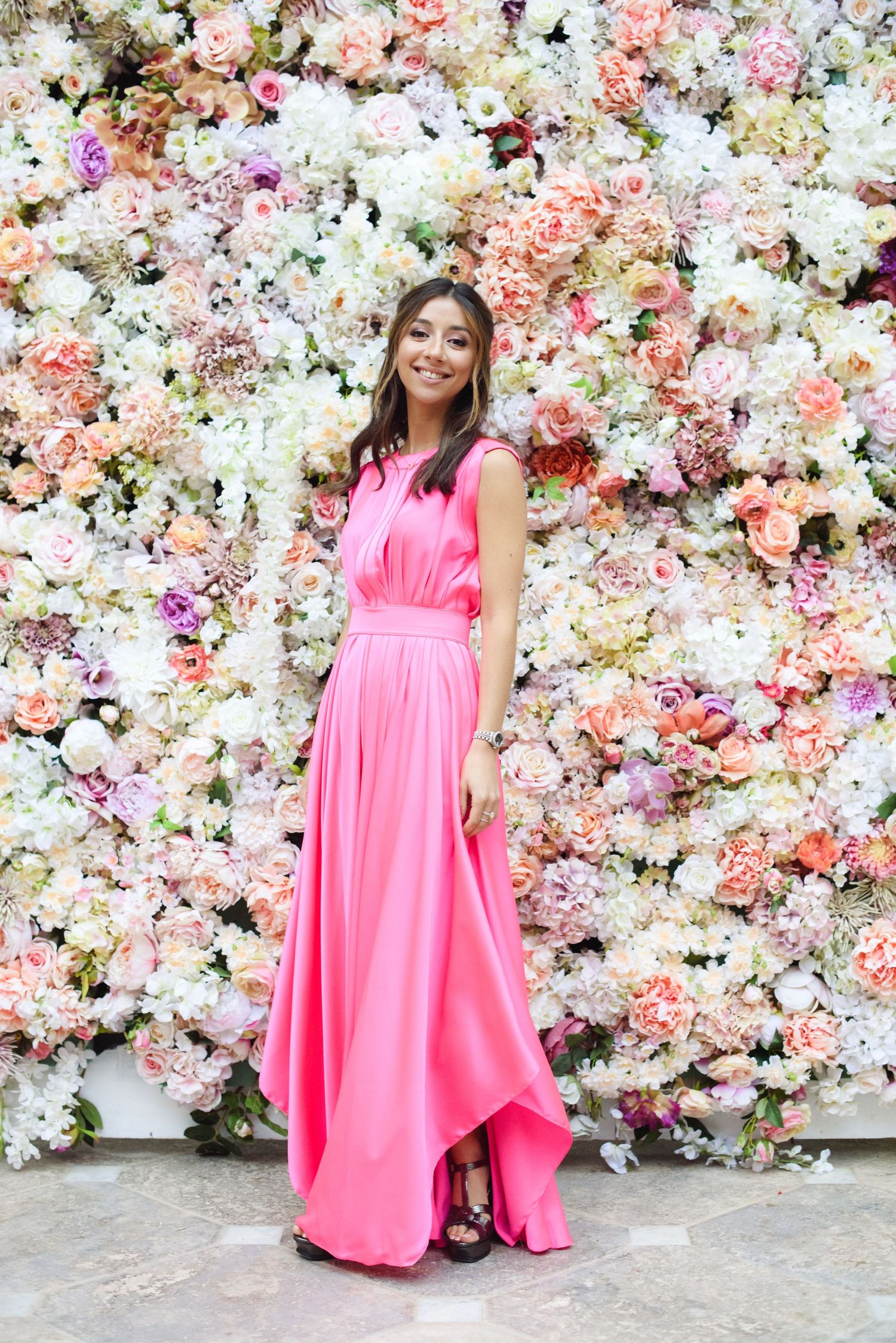Smiling woman in a long pink satin gown stands in front of a lush wall of pink, white and peach flowers.