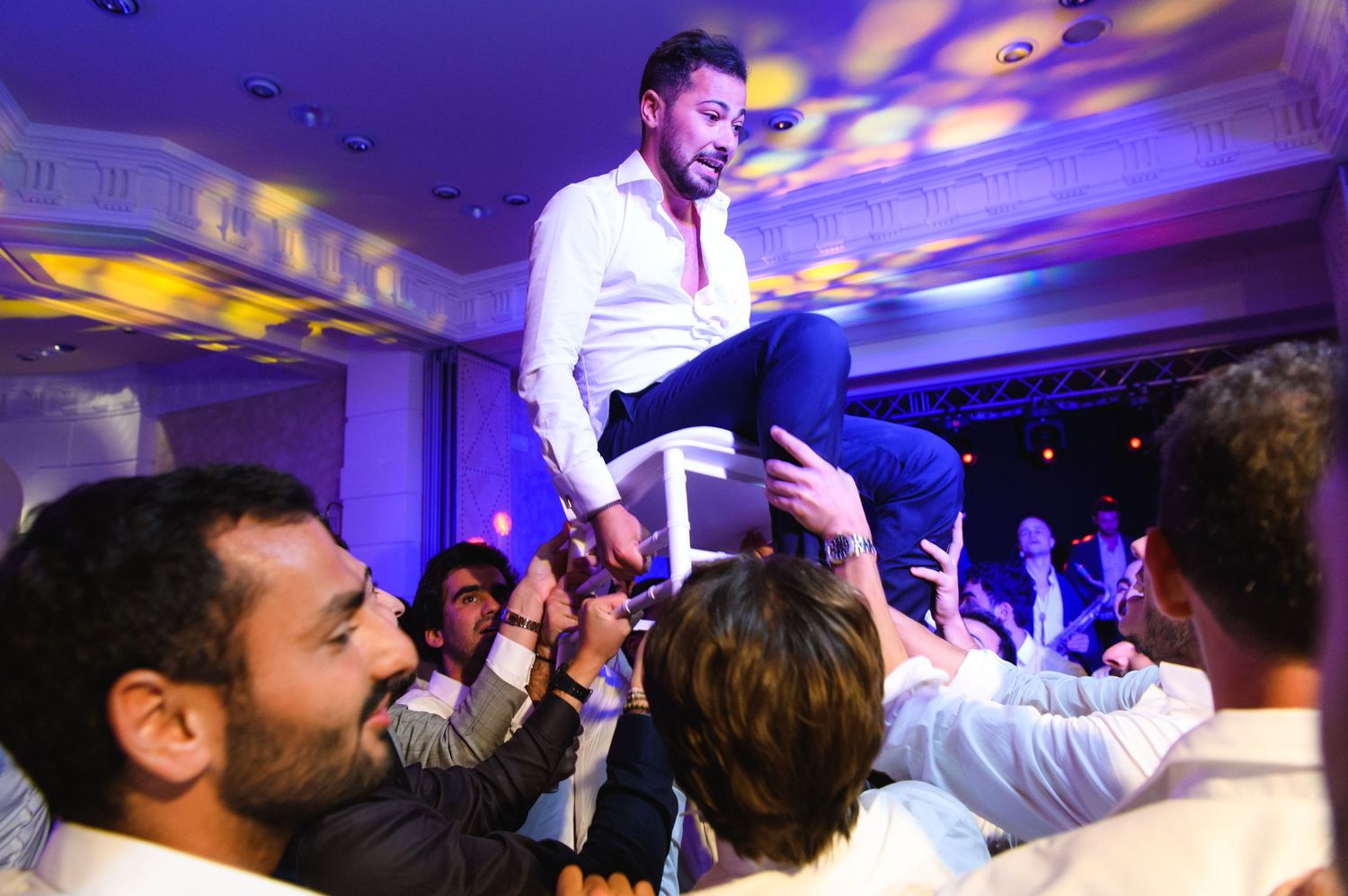Groom in a white shirt sits on a chair raised high by cheering guests during a festive wedding reception celebration.