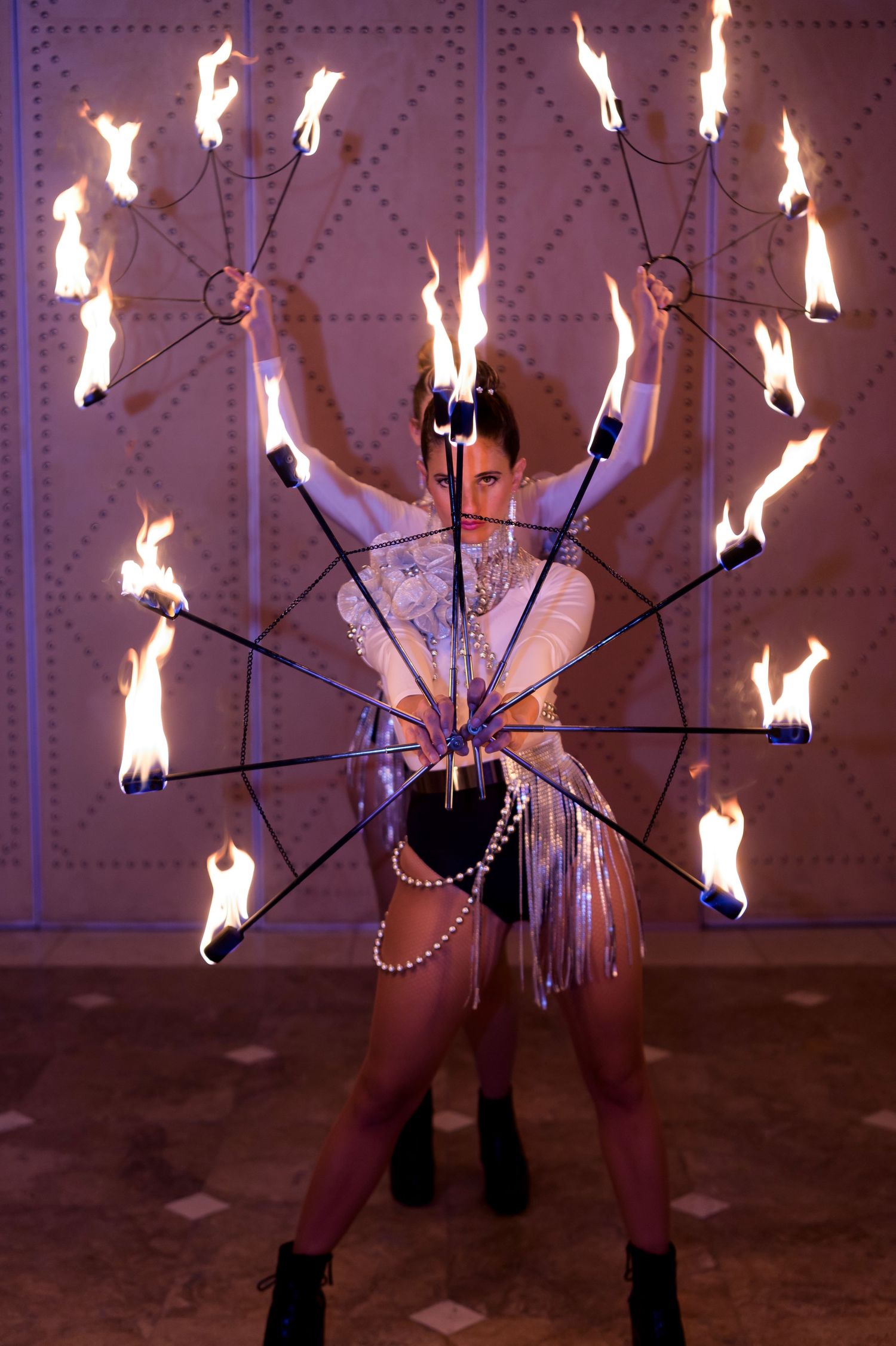 Fire performer in a silver beaded costume spins a circular rig of flaming torches, creating a dramatic display at an upscale event.