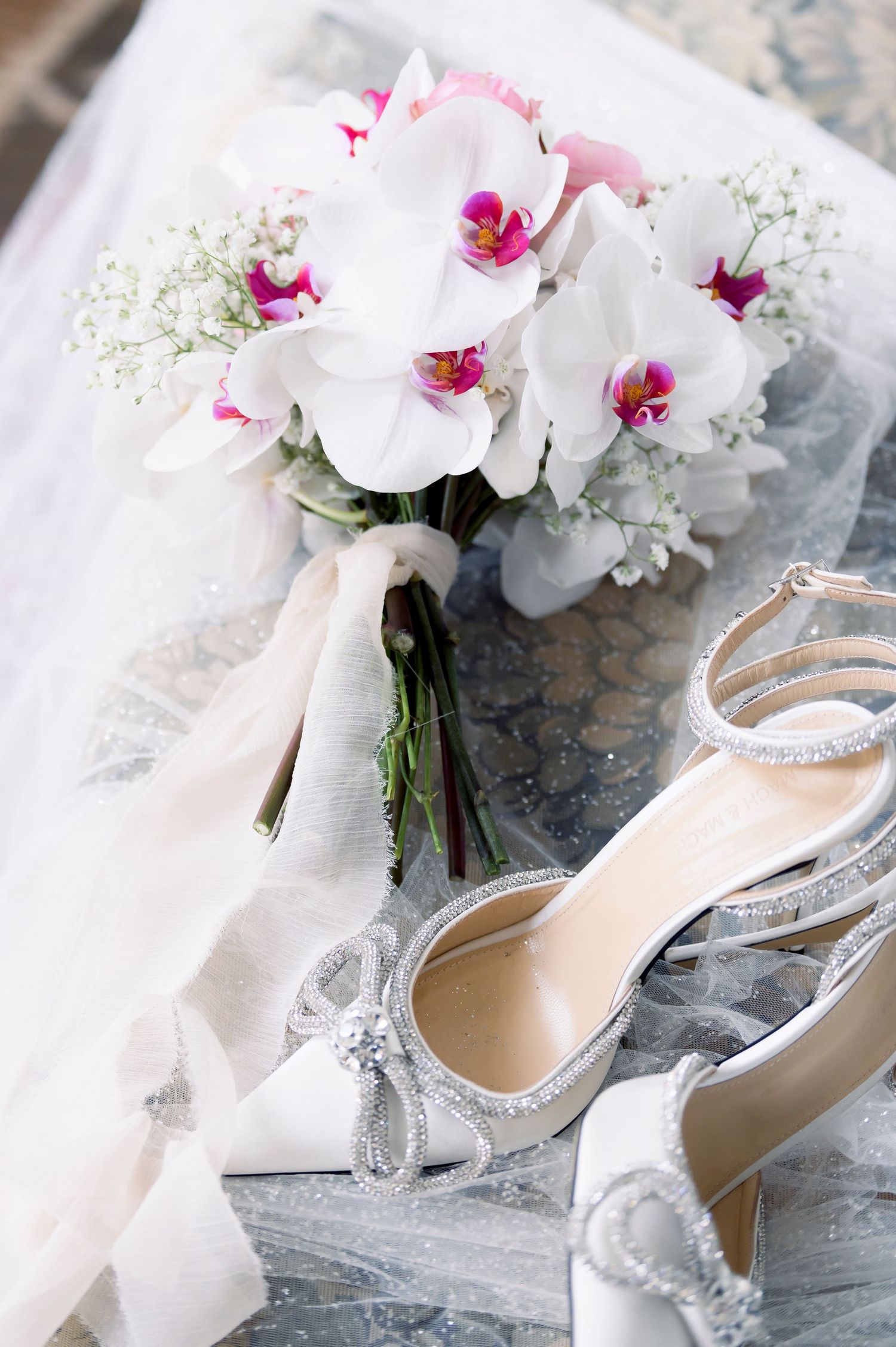 White orchid and baby's breath bridal bouquet with ribbon beside sparkly crystal-trimmed ivory wedding heels on a sheer veil.