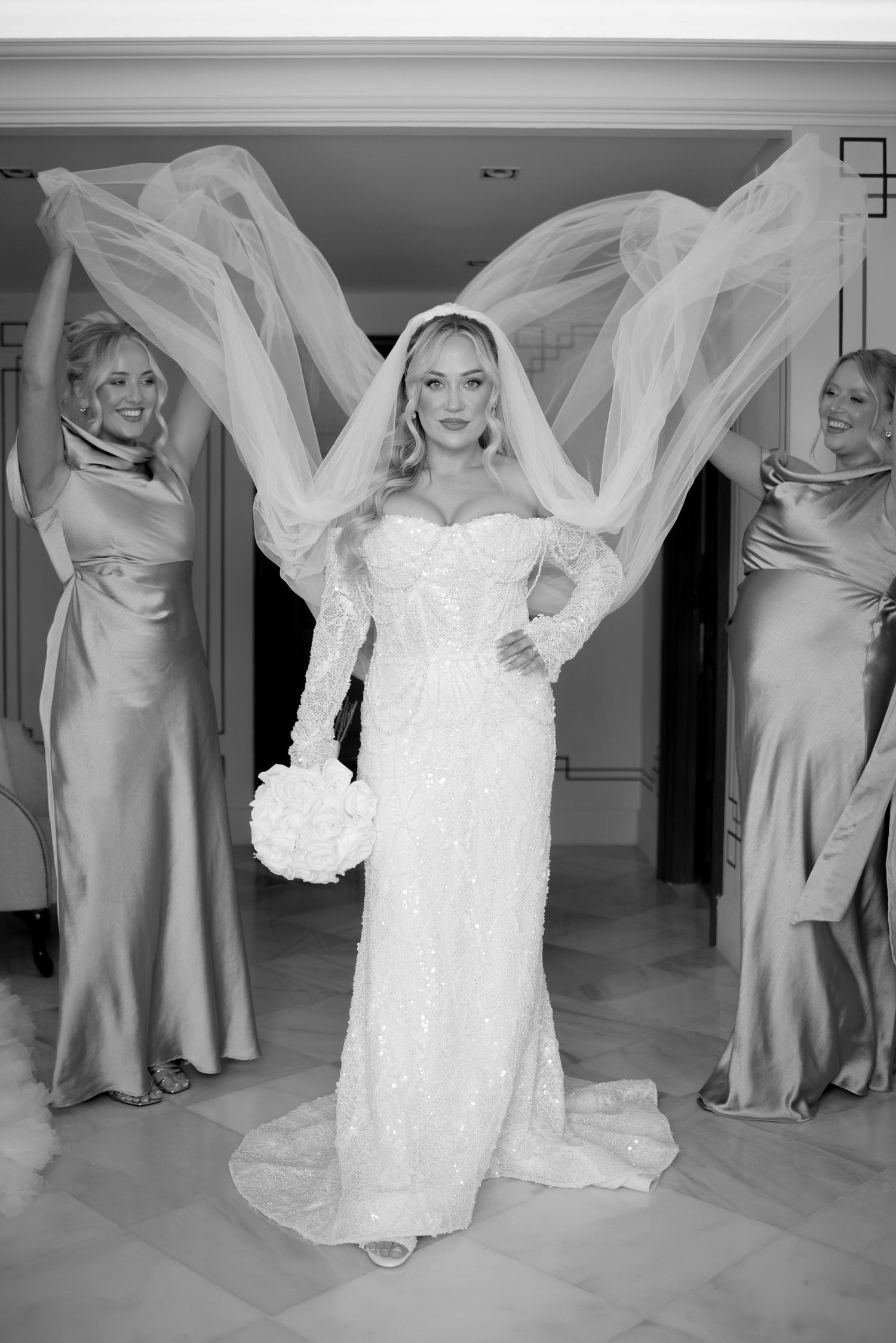 Black and white photo of a bride in a lace wedding gown holding a bouquet as two bridesmaids in satin dresses lift her flowing veil.