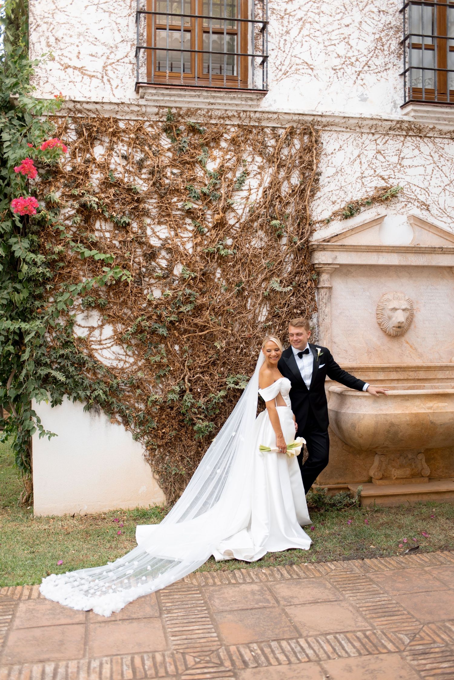 Bride in a white off-shoulder gown with a long lace veil stands beside a groom in a black tuxedo by an ivy-covered wall and stone fountain.