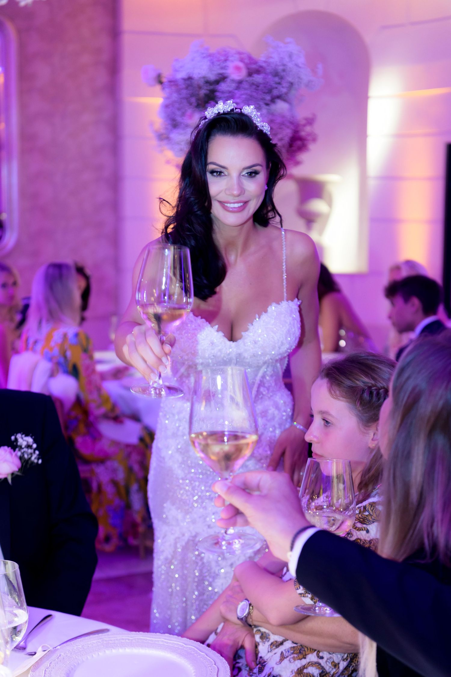 Smiling bride in a sparkling beaded gown raises a glass of white wine to toast guests at an elegant wedding reception.