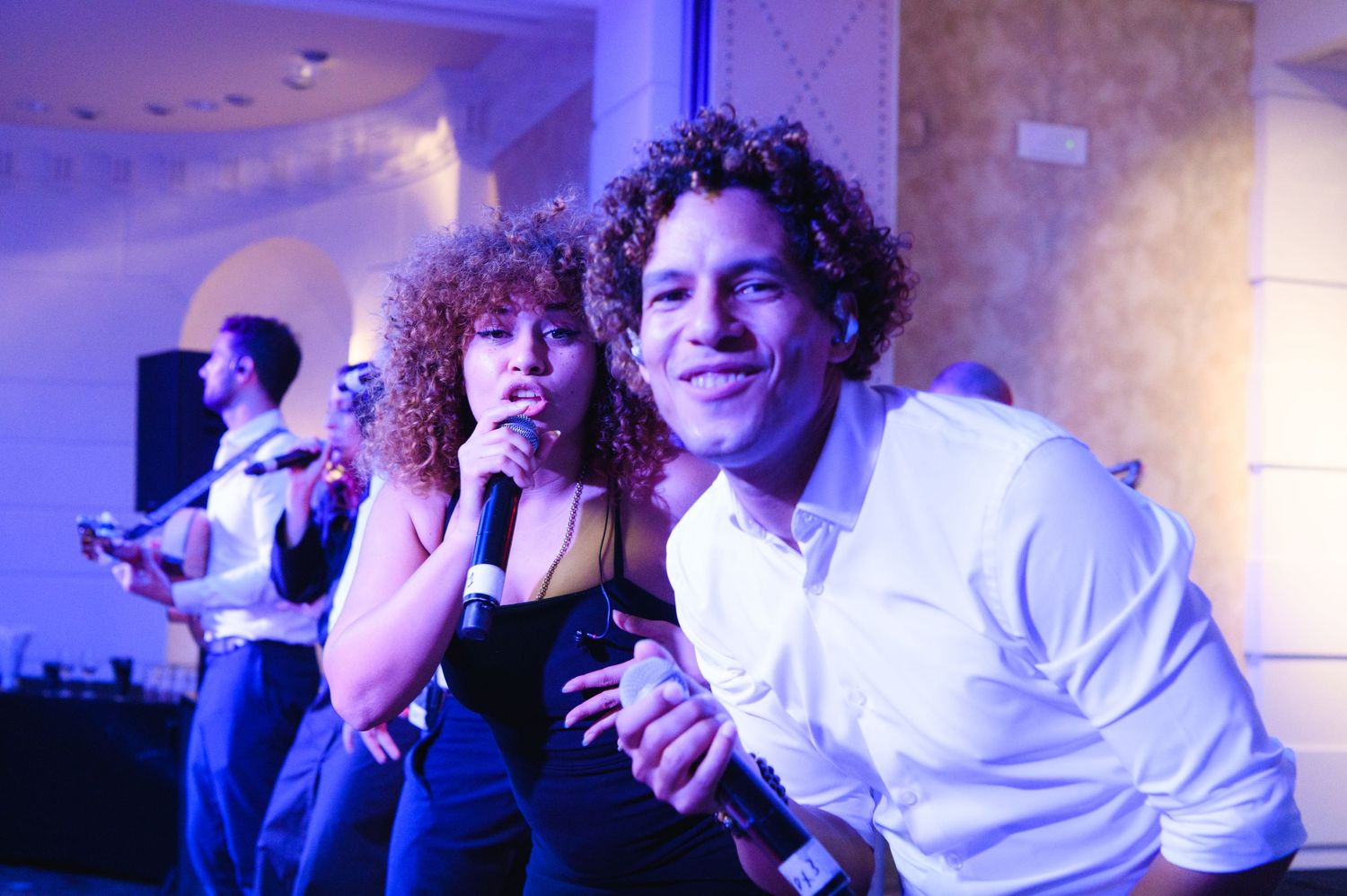 A curly-haired woman sings into a microphone beside a smiling man in a white shirt while a live band performs at a wedding reception.