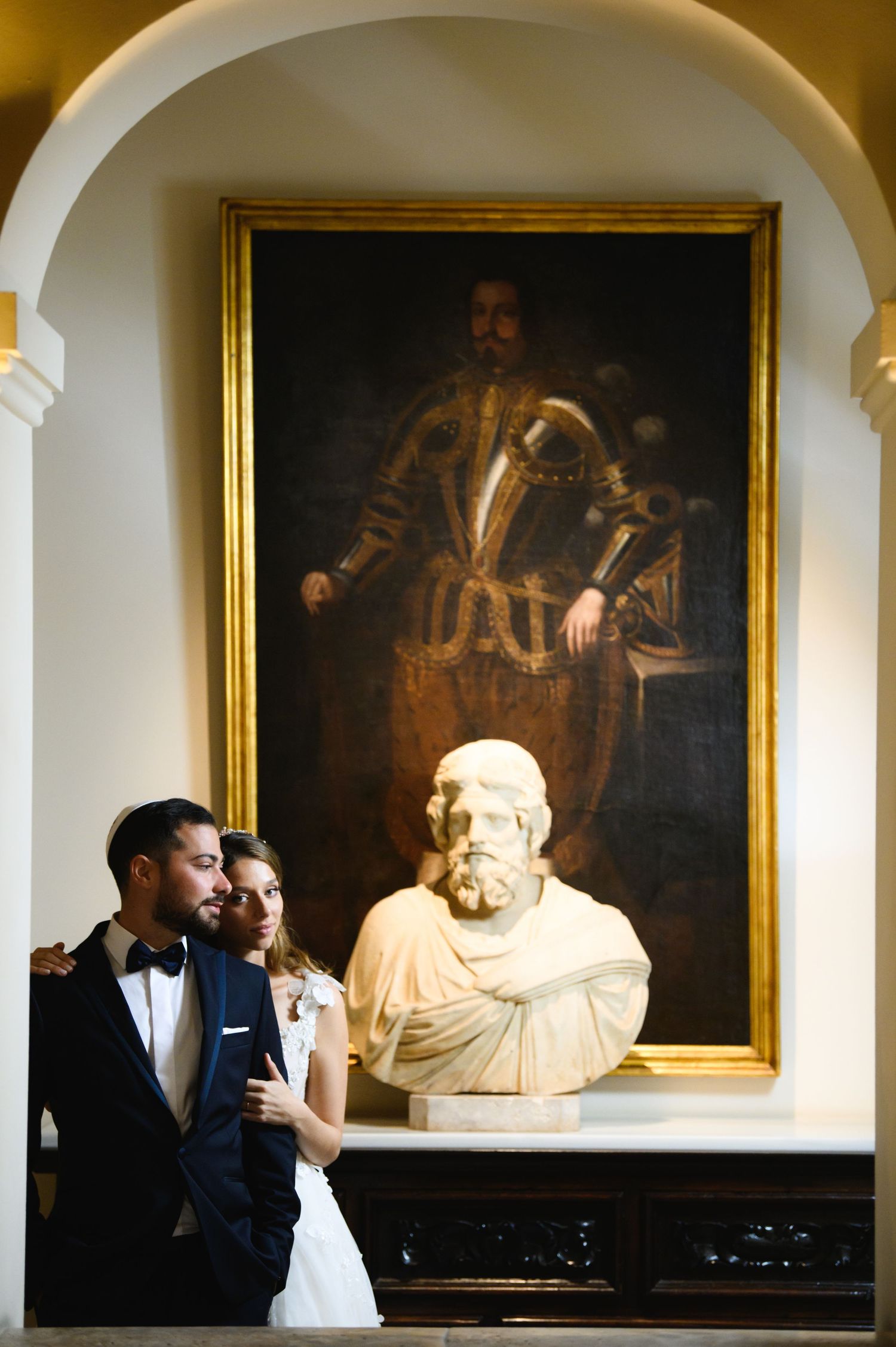 Bride and groom in formal wedding attire pose together beneath an arch beside a marble bust and a large framed portrait.