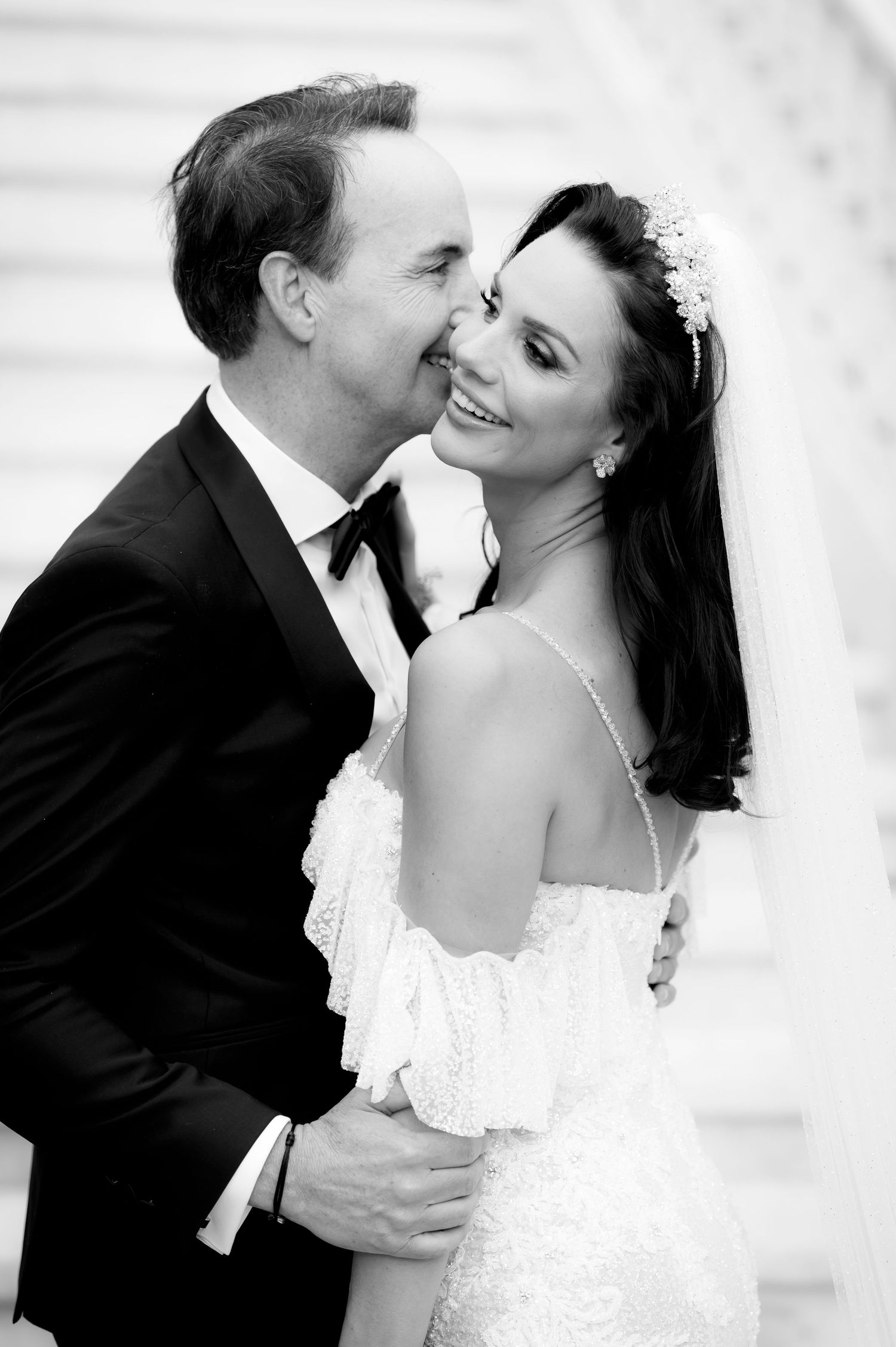 Black and white portrait of a groom in a tuxedo kissing the cheek of a smiling bride wearing a jeweled headpiece and veil.