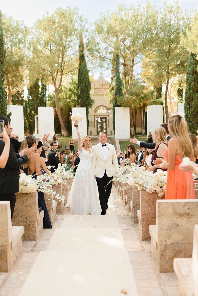 Smiling bride and groom in a white dinner jacket walk down a flower-lined aisle as guests cheer at an outdoor ceremony among cypress trees.