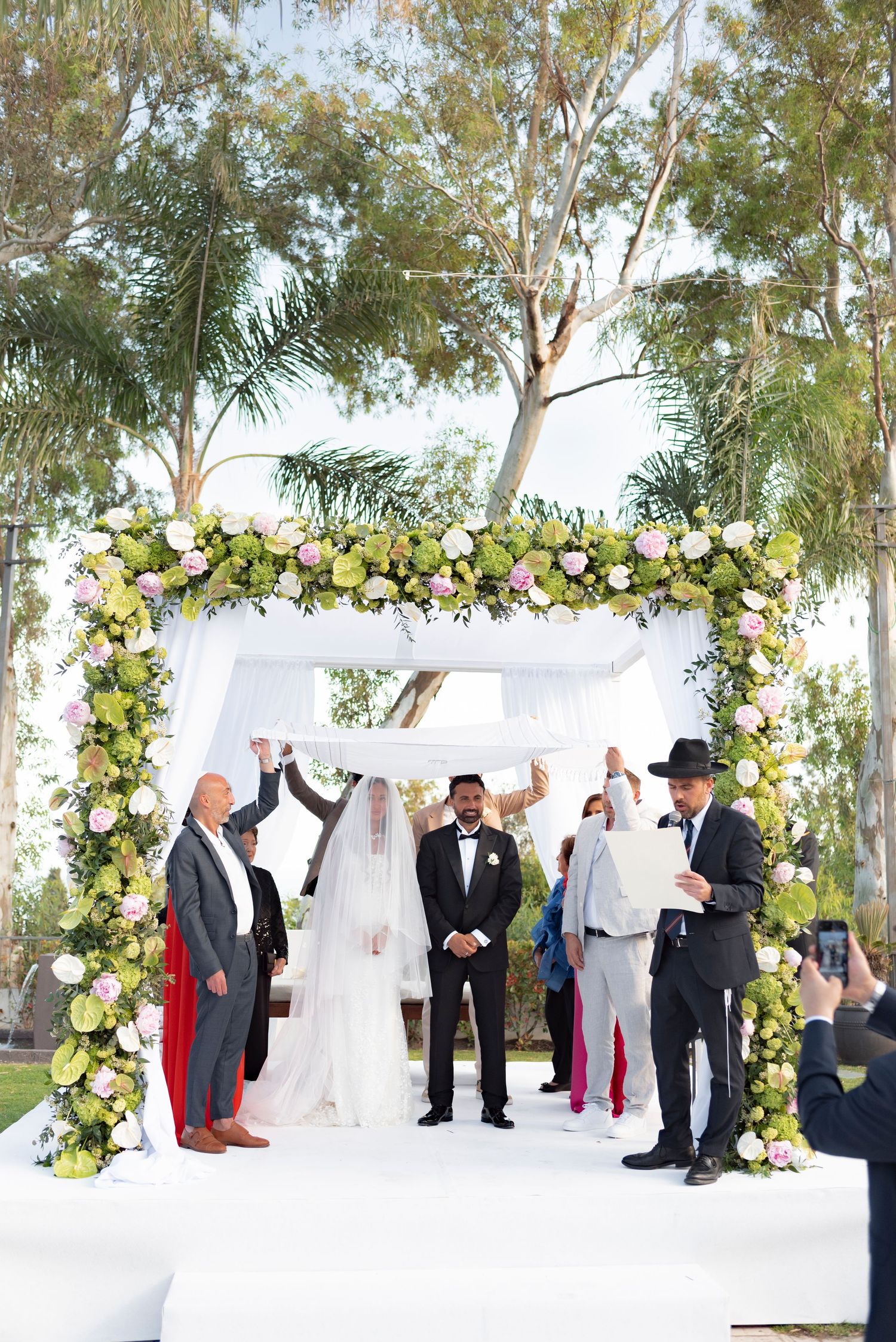 Bride and groom stand under a white chuppah adorned with green and pink florals during an outdoor Jewish wedding ceremony.