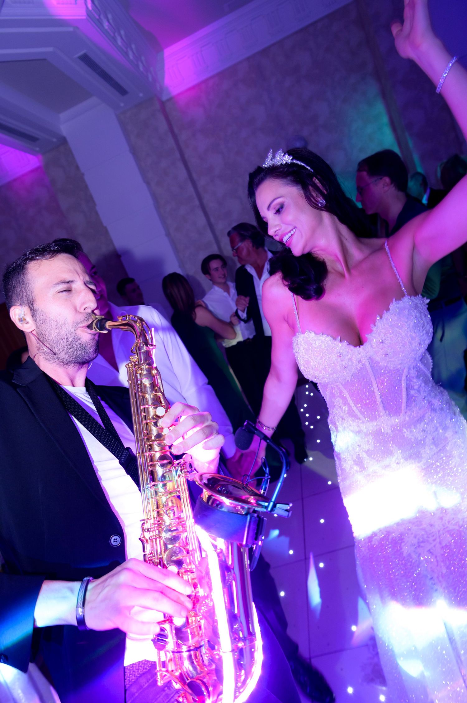 Saxophonist plays beside a smiling bride in a sparkling lace gown and tiara as she dances at a lively wedding reception.