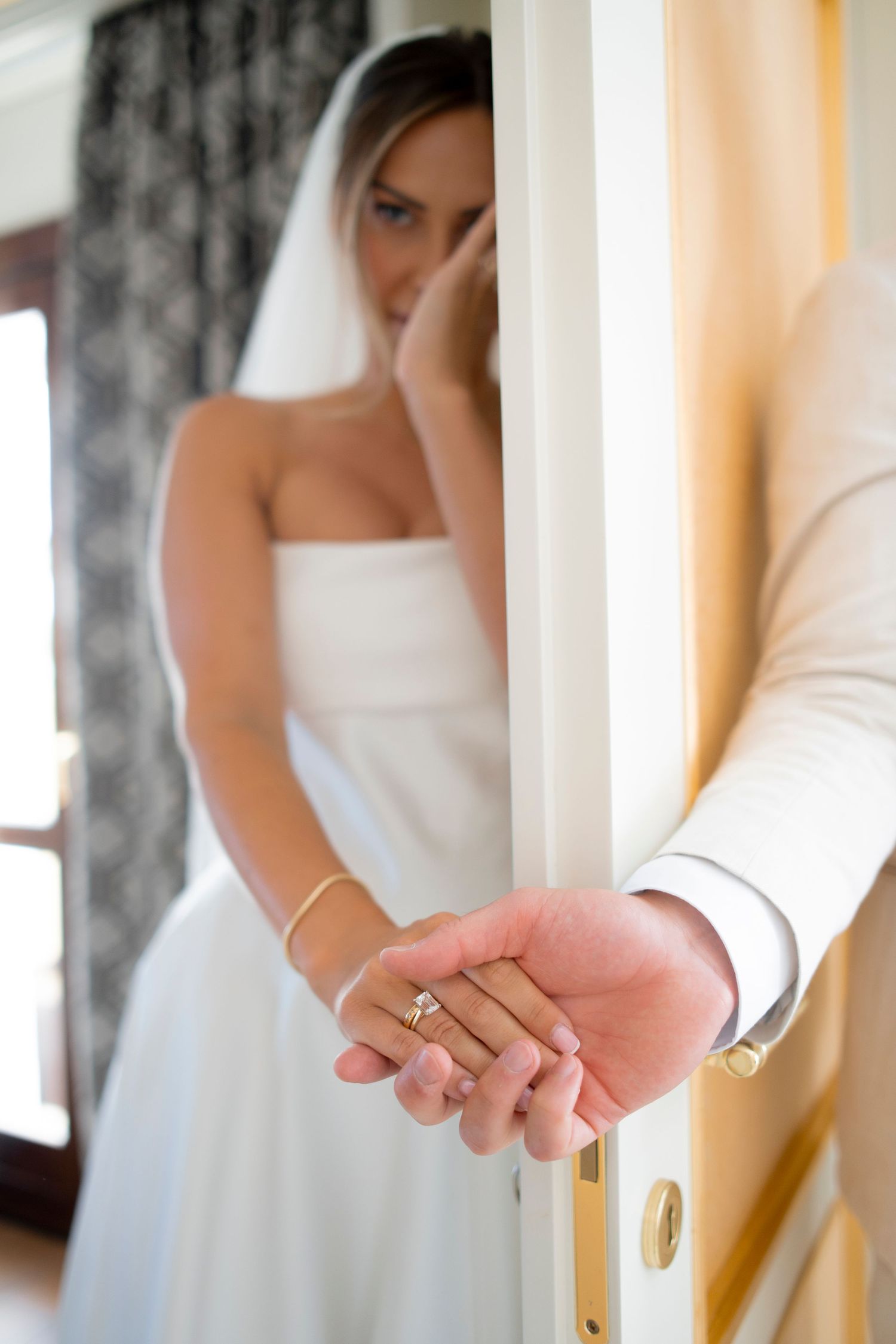 Bride in a white gown and veil holds her groom's hand through a doorway during a first touch moment before their wedding.