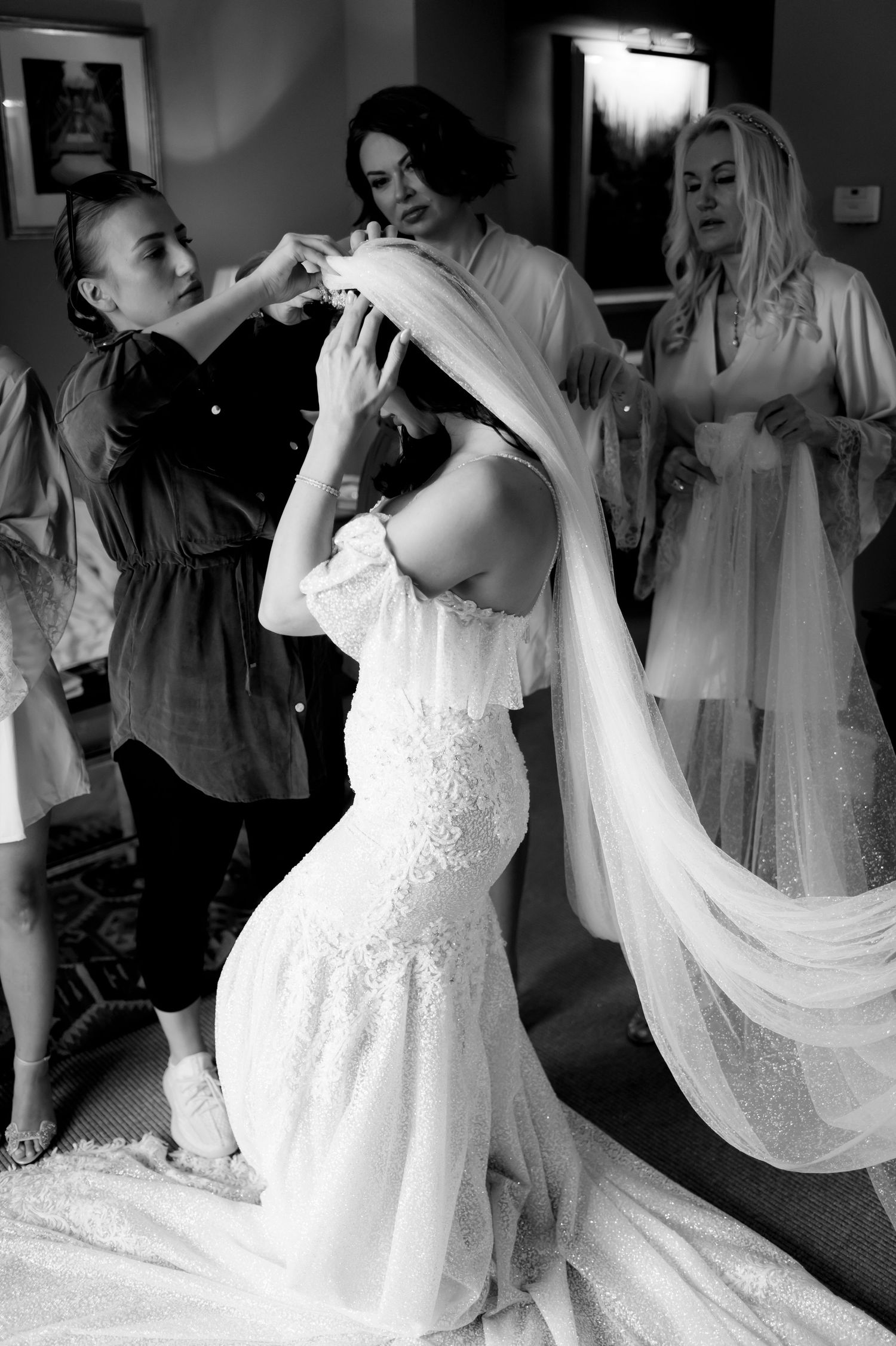 Bride in a lace wedding gown kneels while bridesmaids adjust her long veil during getting-ready moments in a black and white photo.
