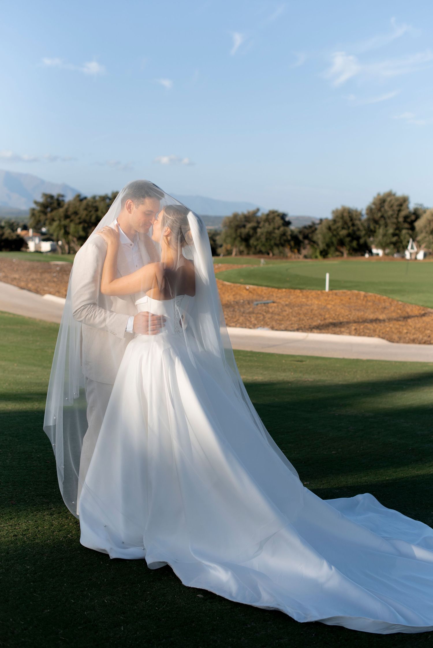 Bride and groom embrace under a sheer veil on a manicured green lawn, with mountains and trees behind them on a sunny wedding day.