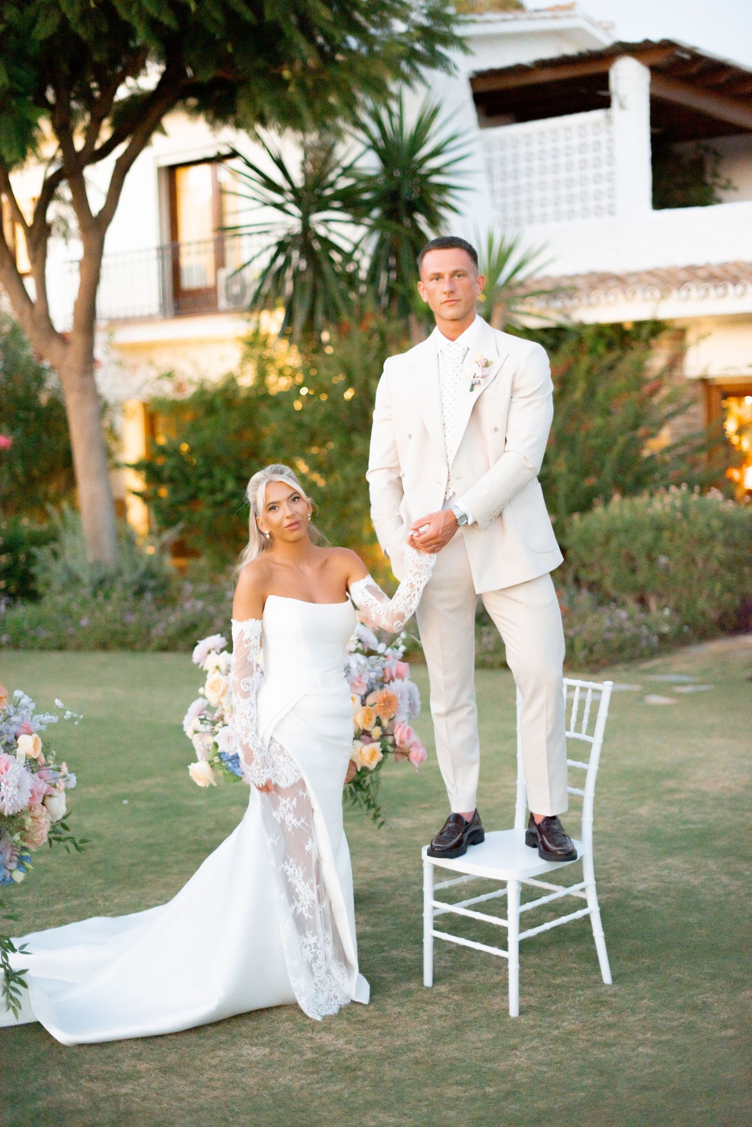 Bride in an off-shoulder white gown with lace sleeves holds a bouquet beside a groom in a cream suit standing on a chair in a garden.