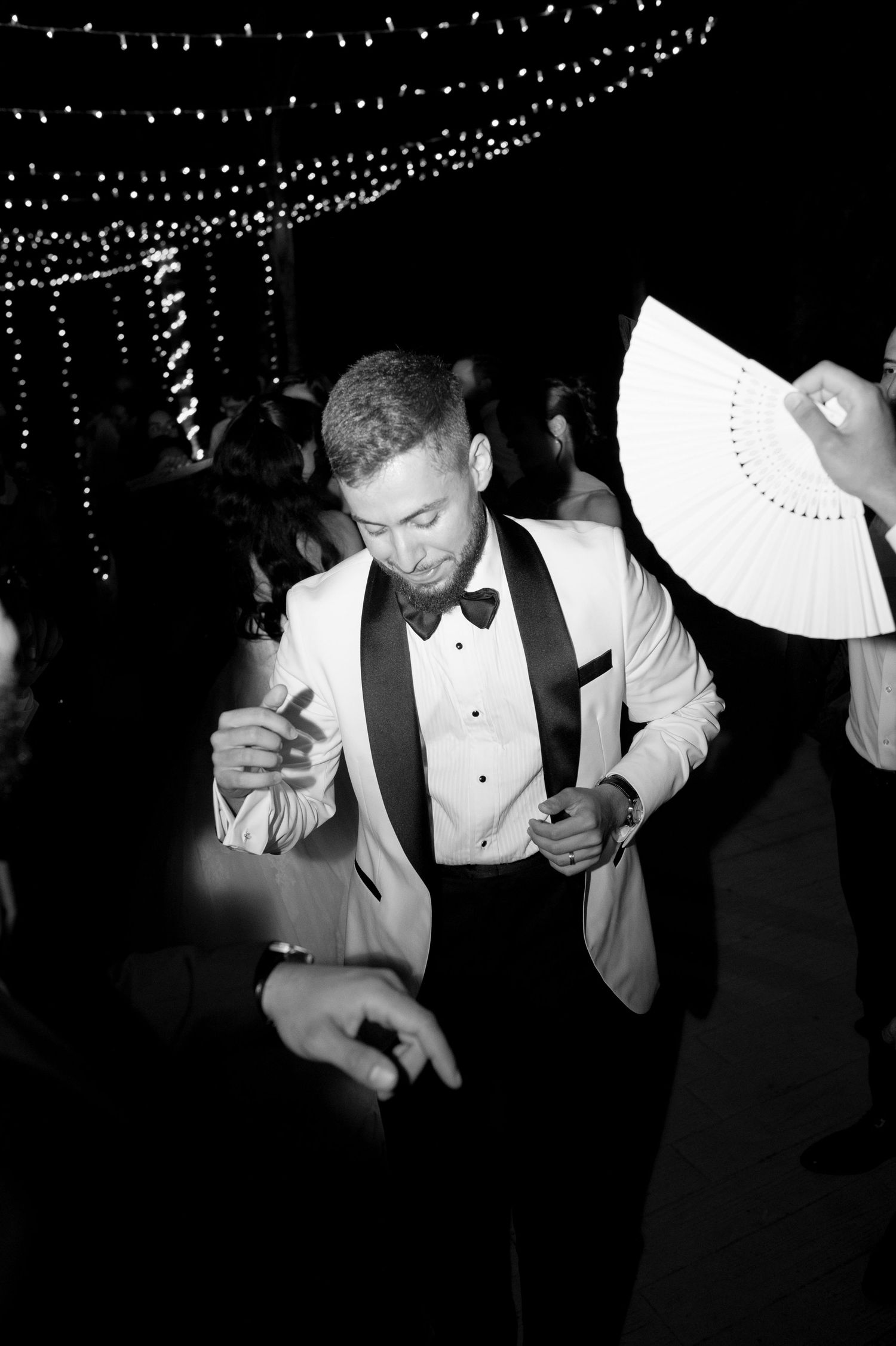 Groom in a white tuxedo jacket and bow tie dances at a nighttime wedding reception under string lights in a black and white photo.