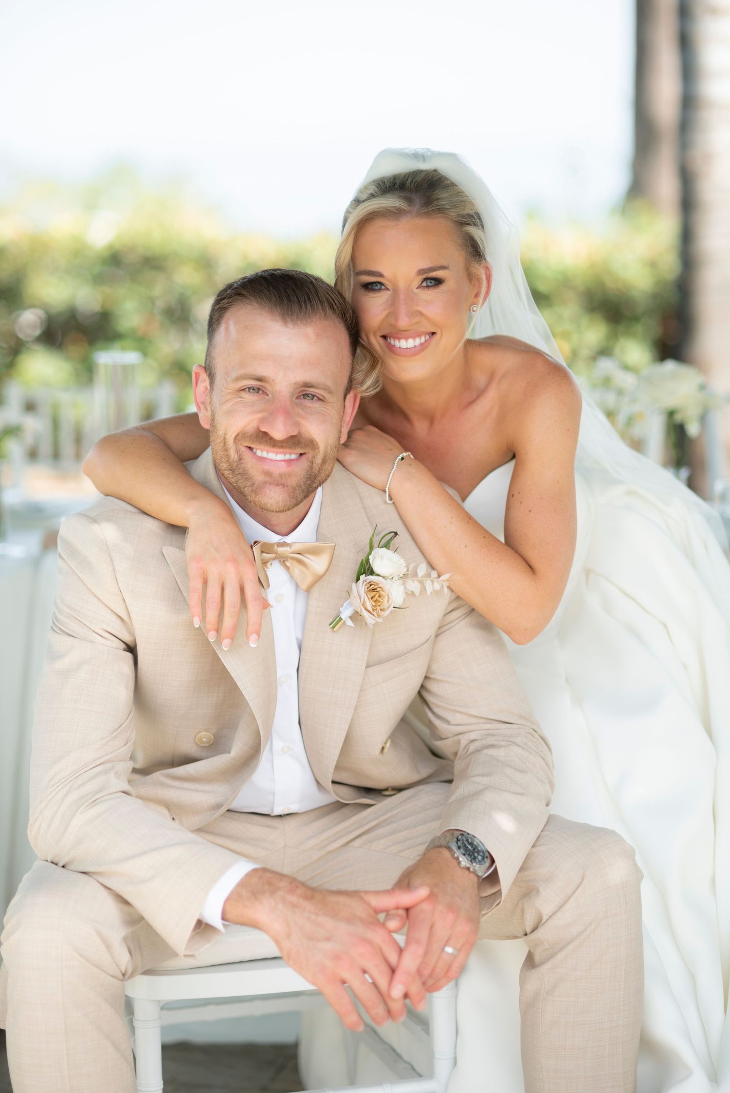 Smiling bride in a white gown and veil embraces her seated groom, who wears a tan suit and bow tie, on their outdoor wedding day.