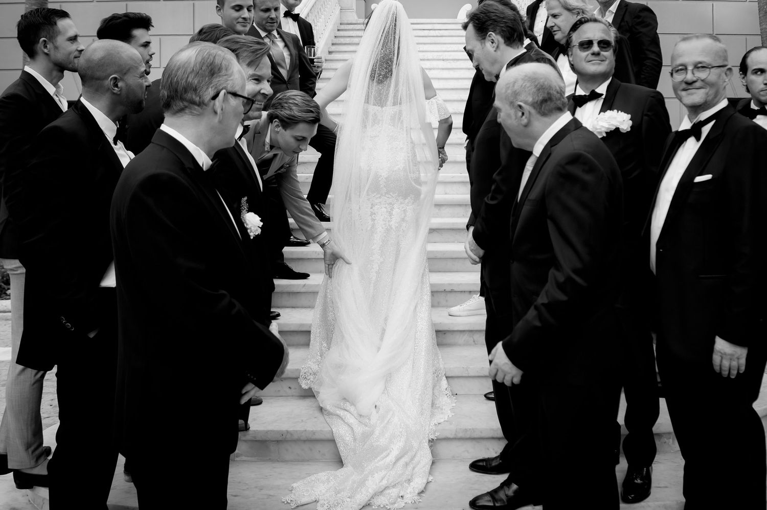 Black and white photo of a bride in a long veil and gown standing on stone steps while formally dressed wedding guests in suits watch.