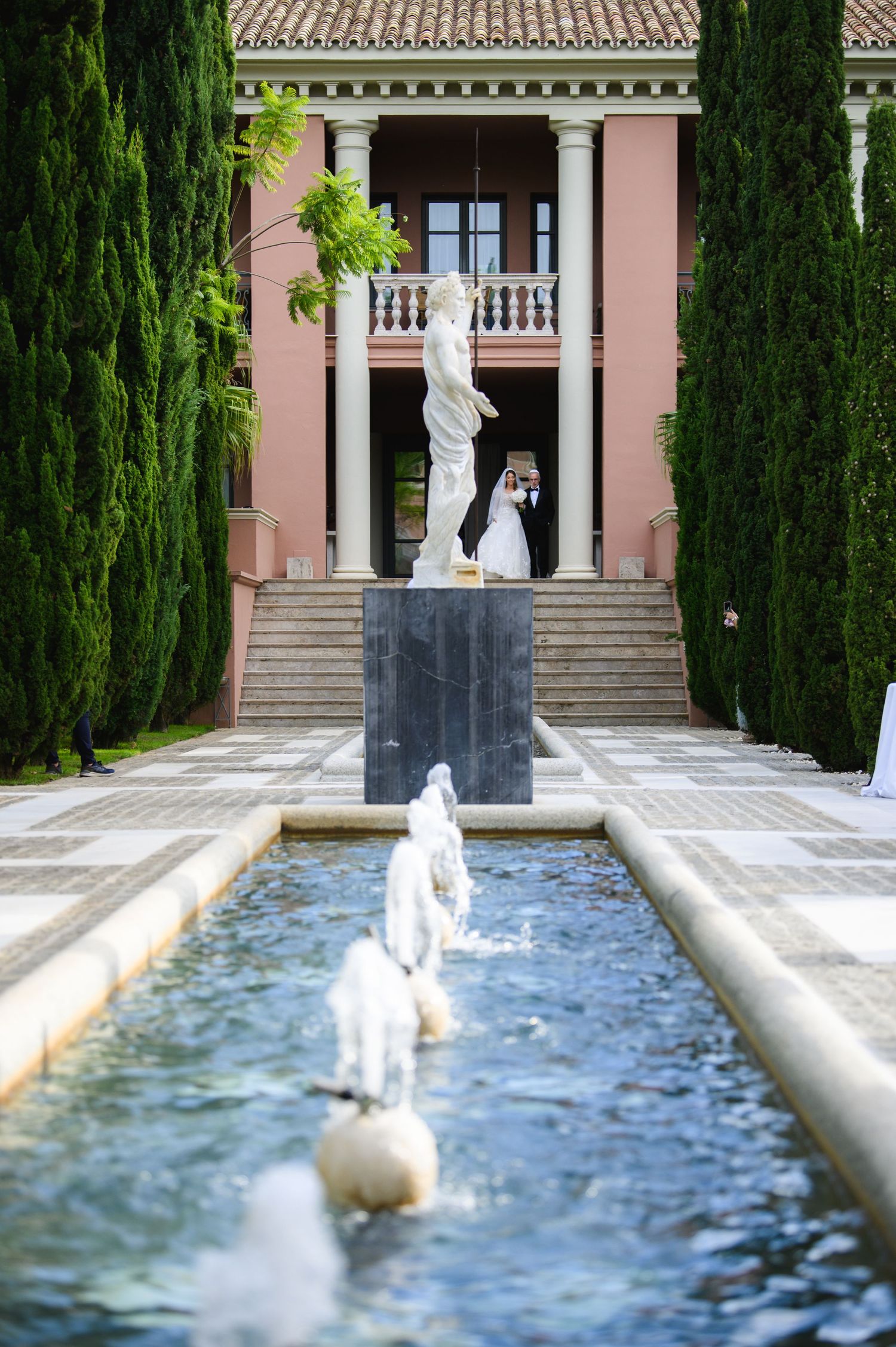 Bride in a white gown stands beyond a marble statue and reflecting pool with fountains at an elegant pink villa with cypress trees.