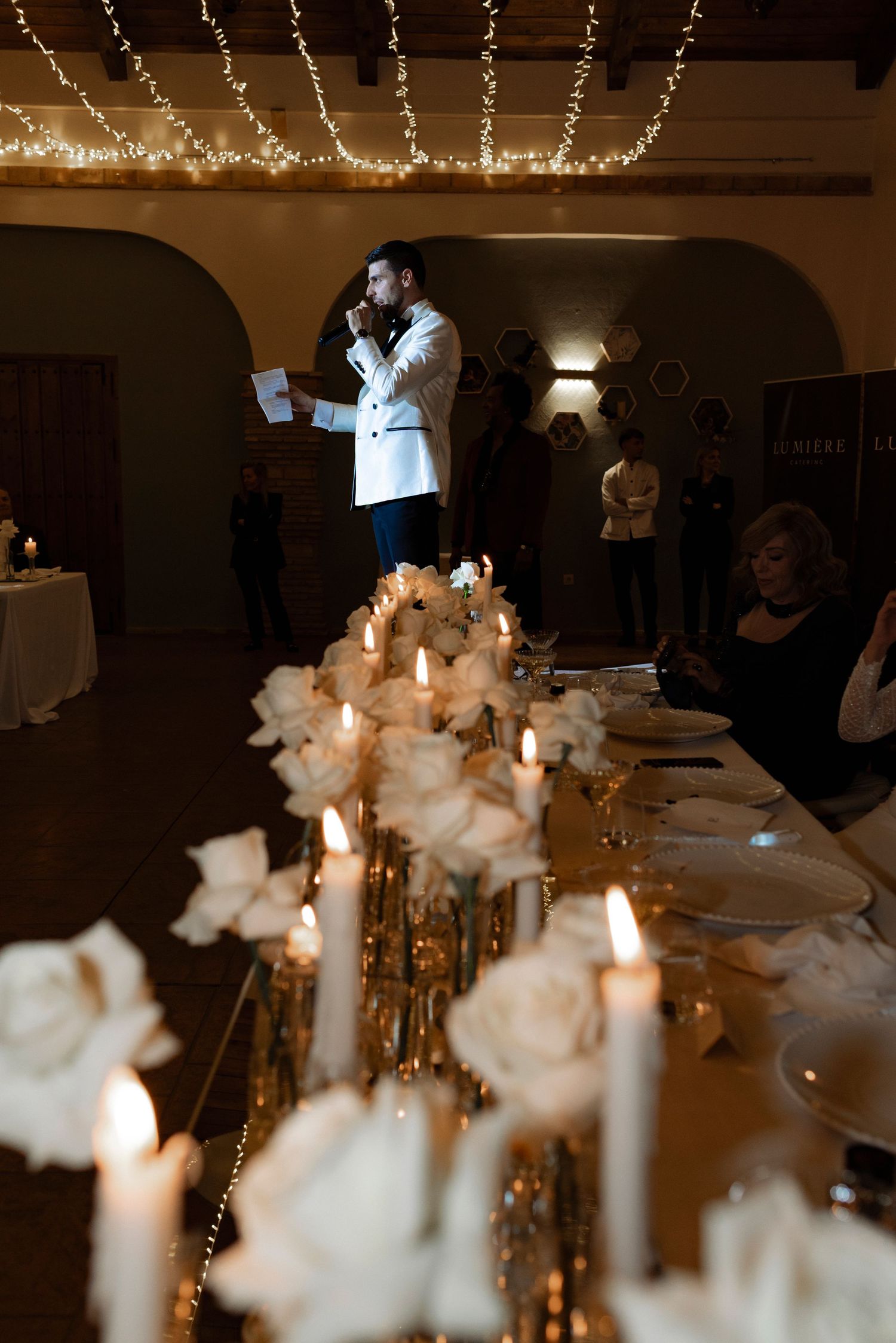 Man in a white jacket speaks from notes beside a candlelit reception table with white roses, as guests look on beneath string lights.