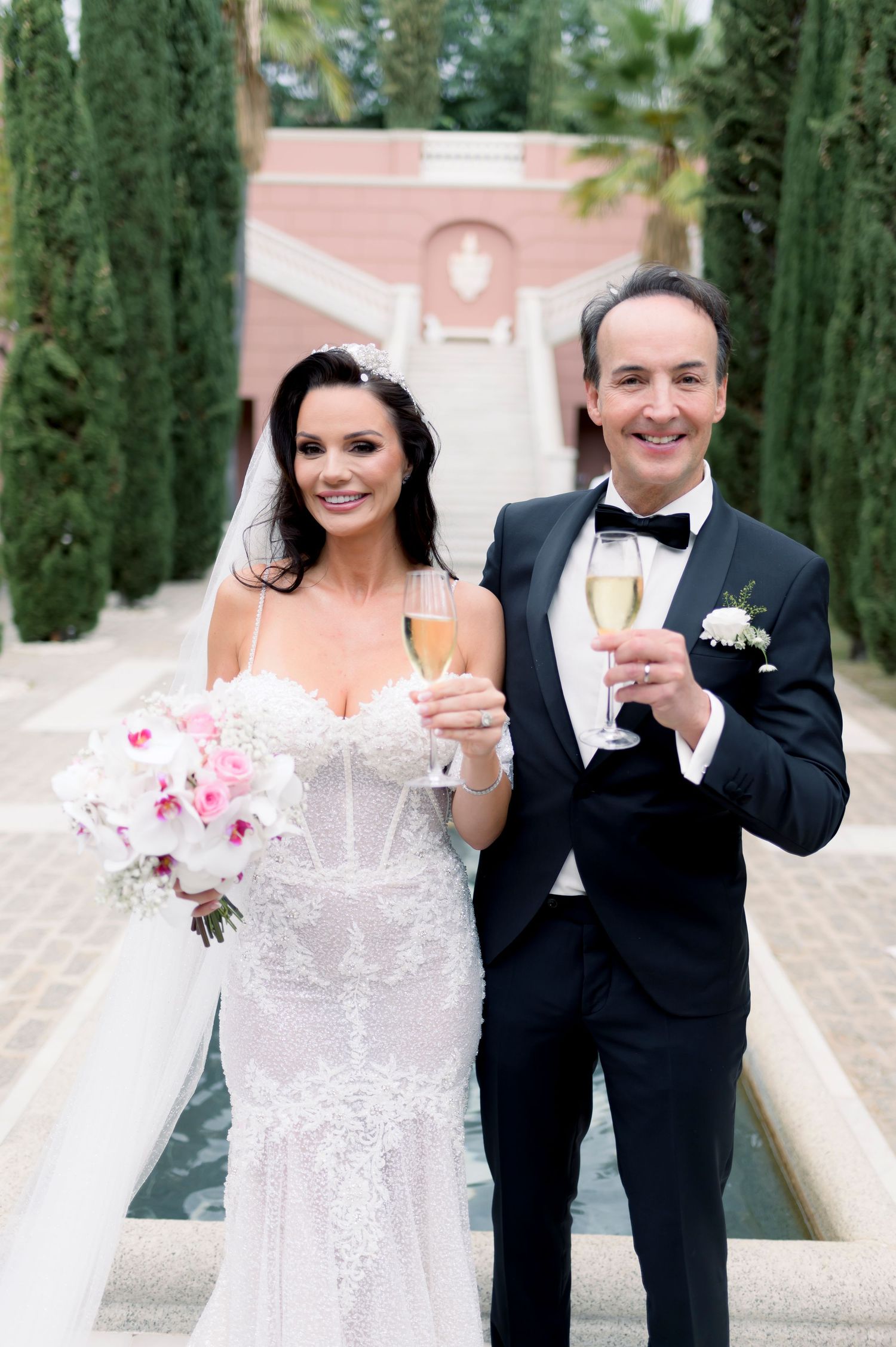 Smiling bride and groom in a black tuxedo toast with champagne flutes by a garden fountain, holding a pink and white bridal bouquet.