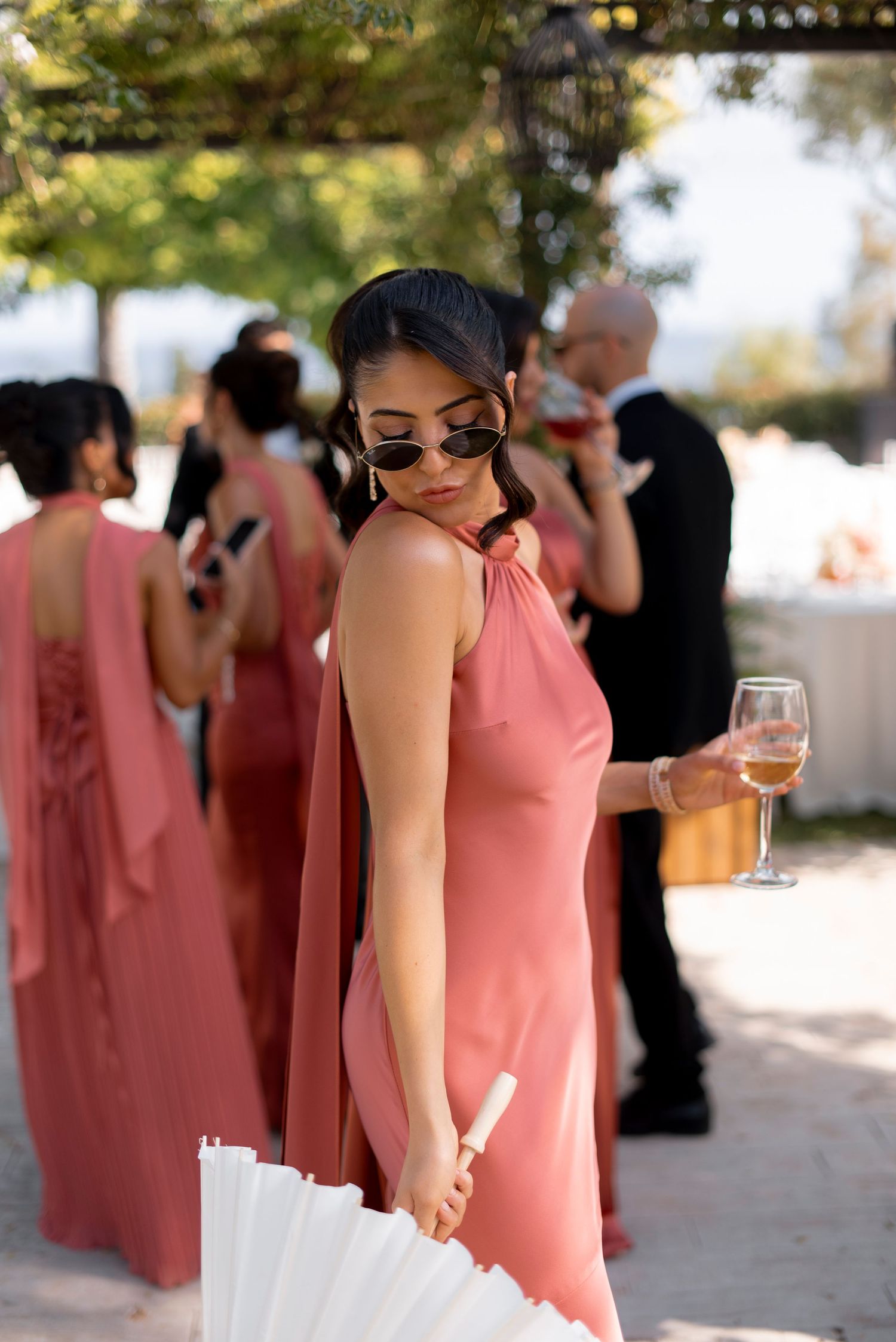 Bridesmaid in a blush pink satin dress and sunglasses holds a wine glass and white folding fan at an outdoor wedding reception.