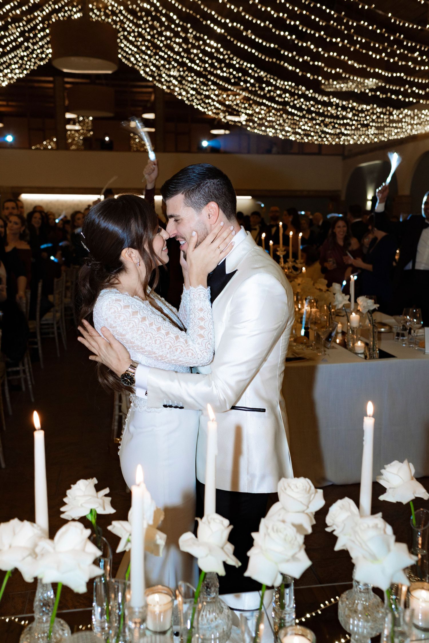 Bride in a white lace gown and groom in a white tuxedo jacket share a tender moment at a candlelit wedding reception with white roses.