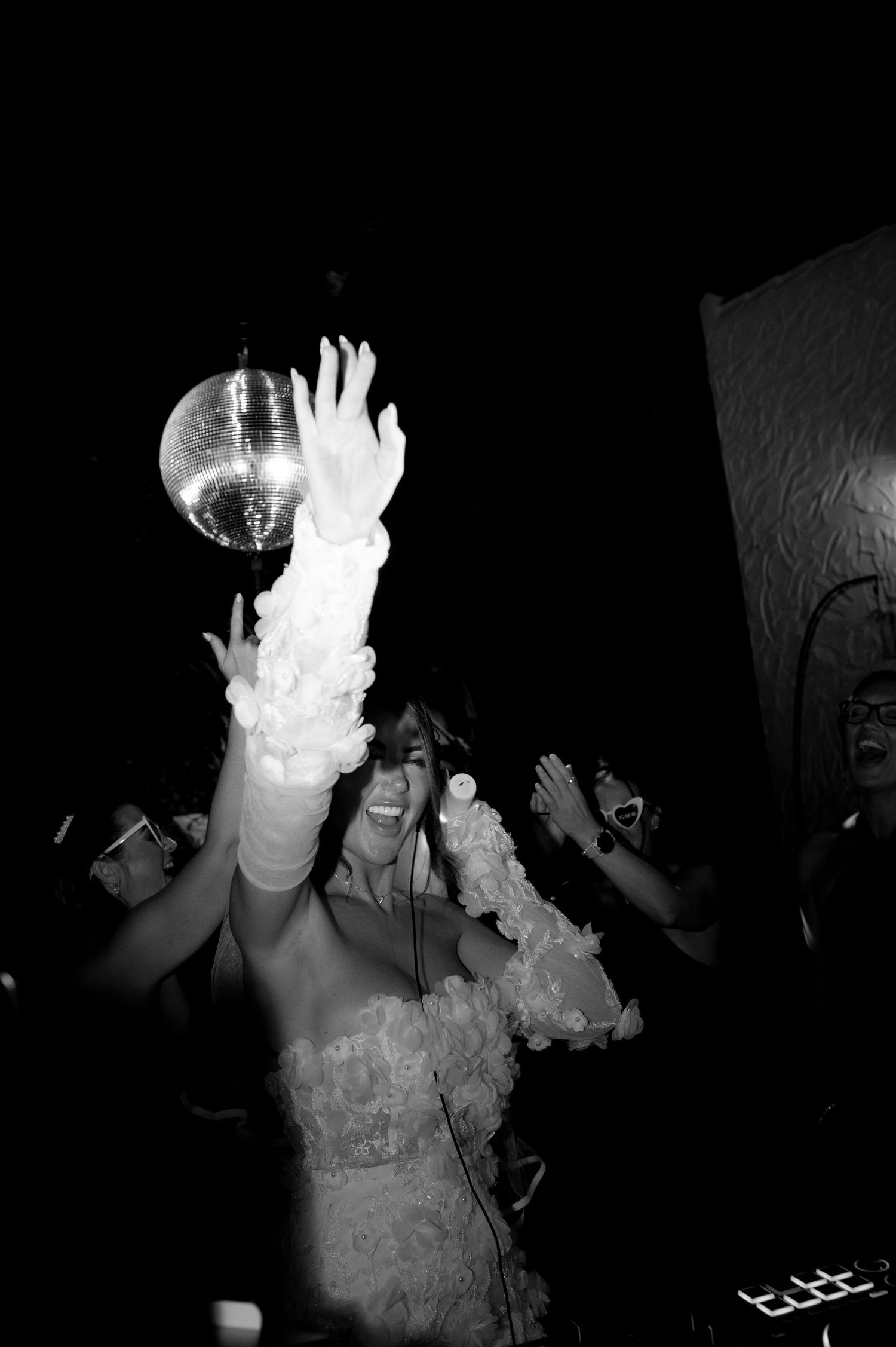 Black and white photo of a smiling bride in a ruffled gown and long gloves dancing with raised arms beneath a disco ball at a wedding party.