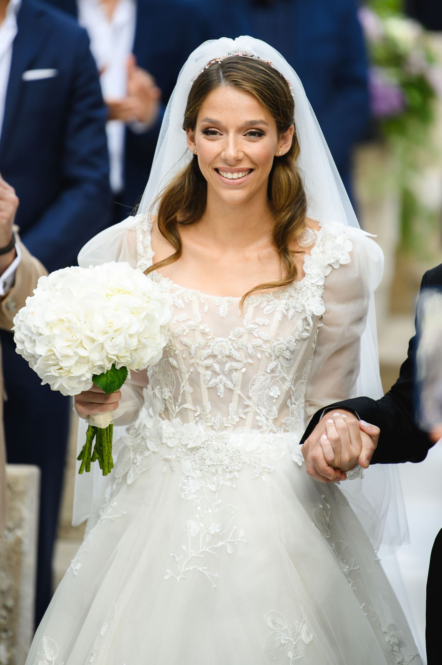 Smiling bride in a lace gown with sheer sleeves and veil holds a white rose bouquet while clasping a hand.