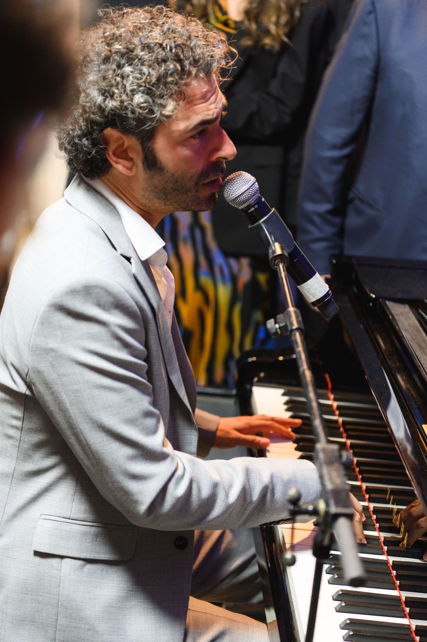 A curly-haired bearded man in a gray blazer sings into a microphone while playing a black grand piano at a live event.