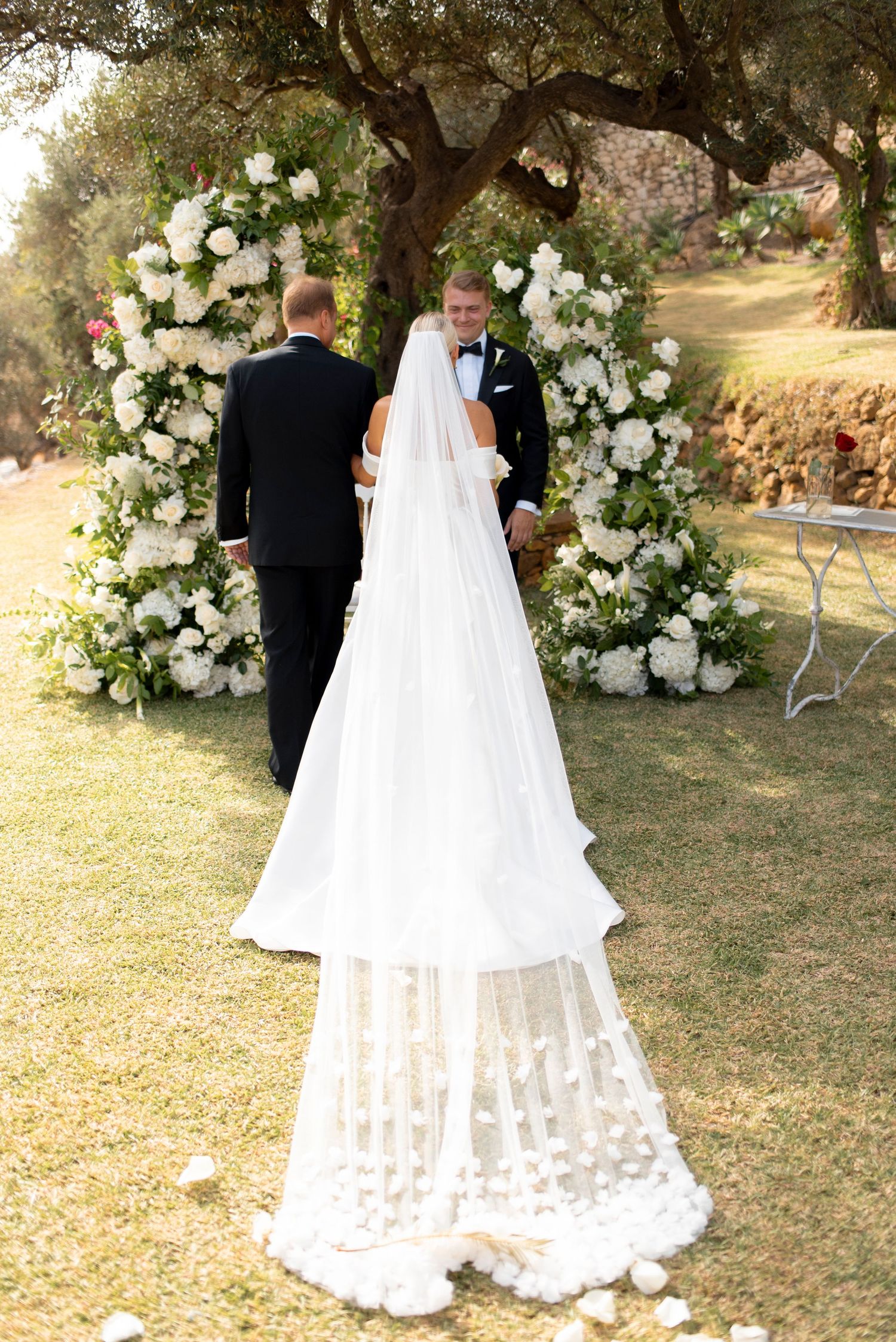 Bride in a long lace-trimmed veil and gown faces two men in dark suits before a white floral arch at an outdoor olive grove wedding.